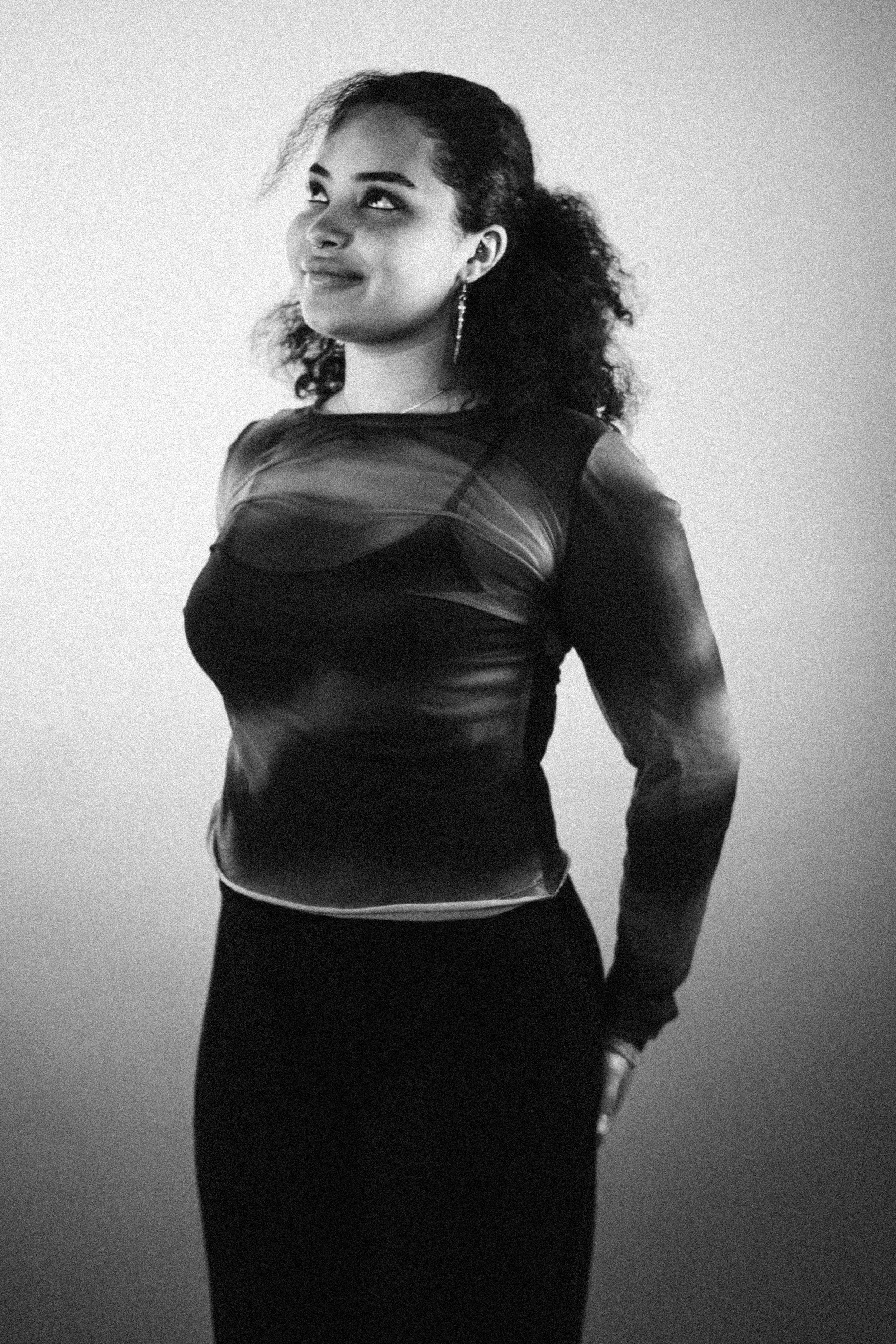 Black and white photo of a woman with curly hair, wearing earrings, a sheer-top, and a long skirt, standing against a plain wall.