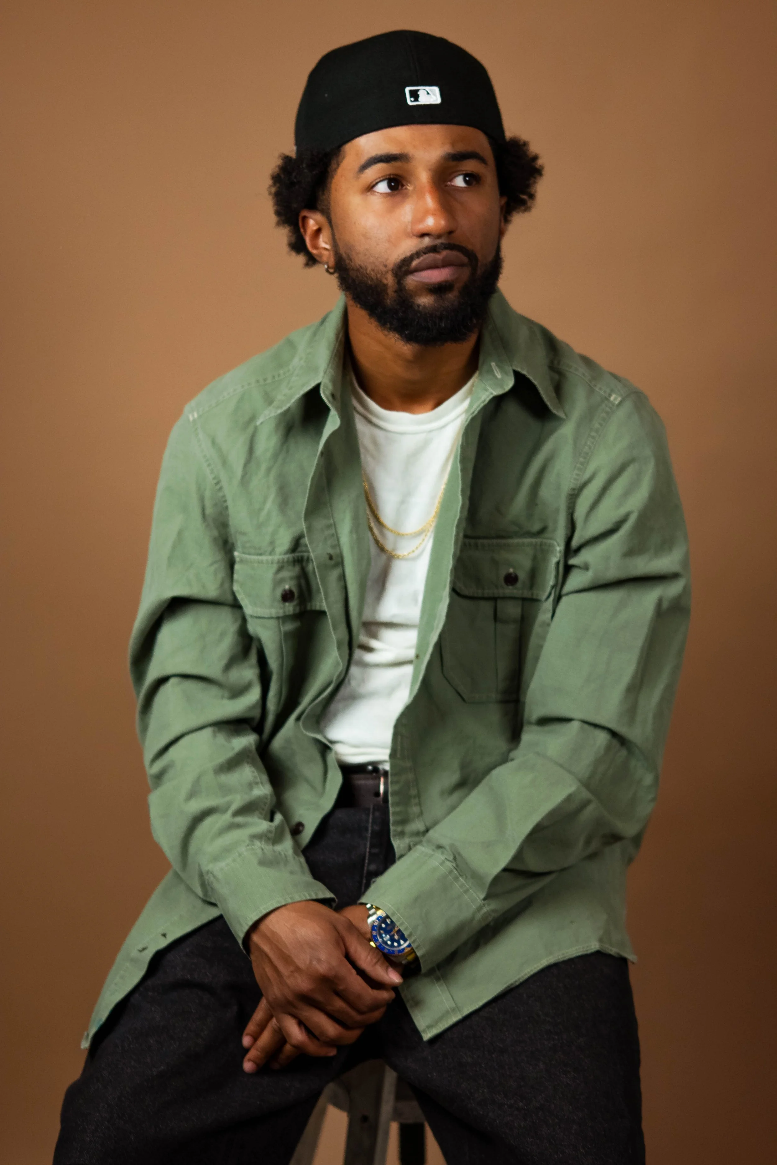 A man with dark skin and a beard, wearing a black cap, green jacket, white t-shirt, gold chains, and a watch, sitting on a stool against a brown background.