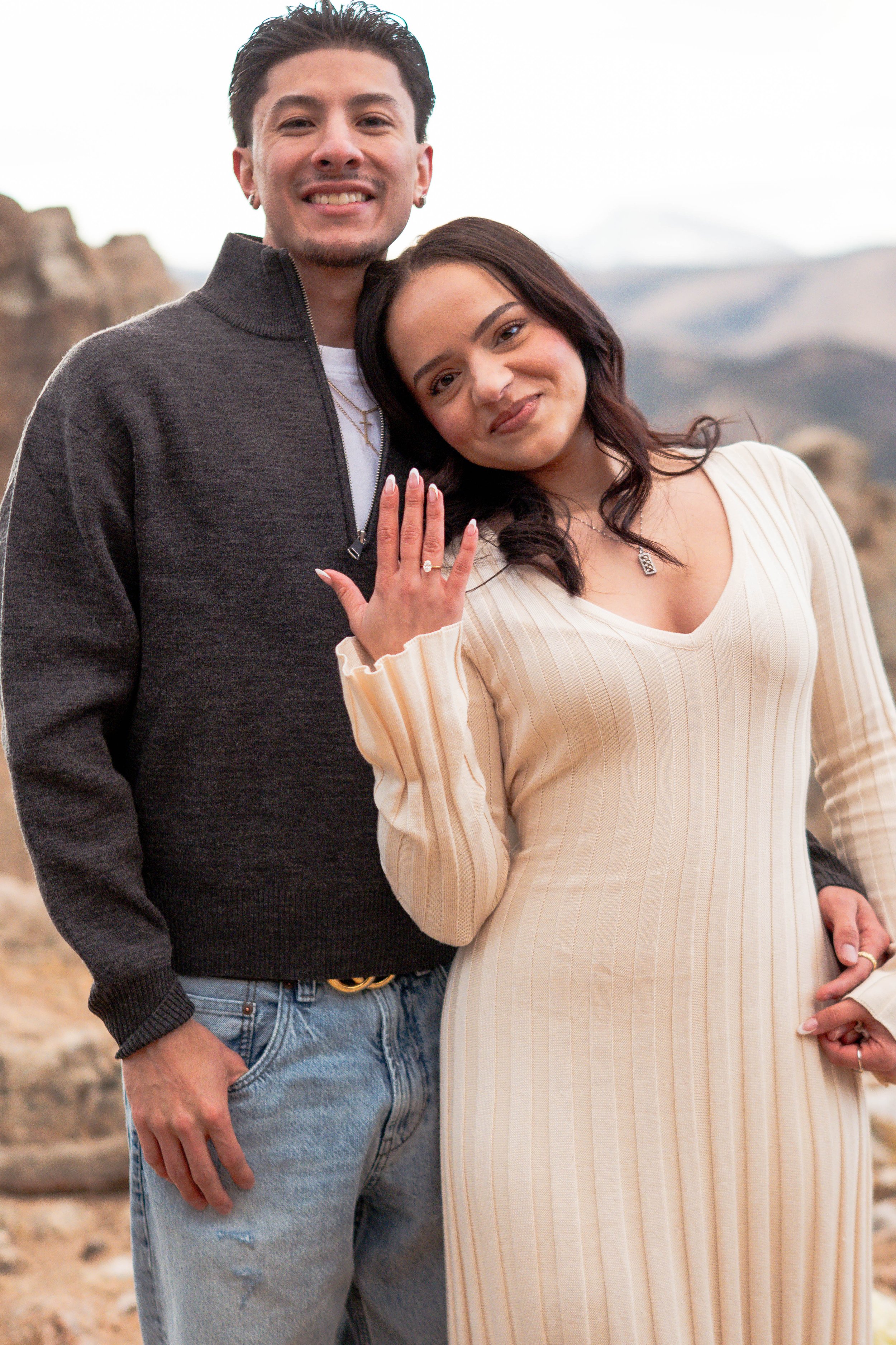 A young couple standing outdoors with mountains in the background; the woman shows an engagement ring on her finger, and both are smiling.