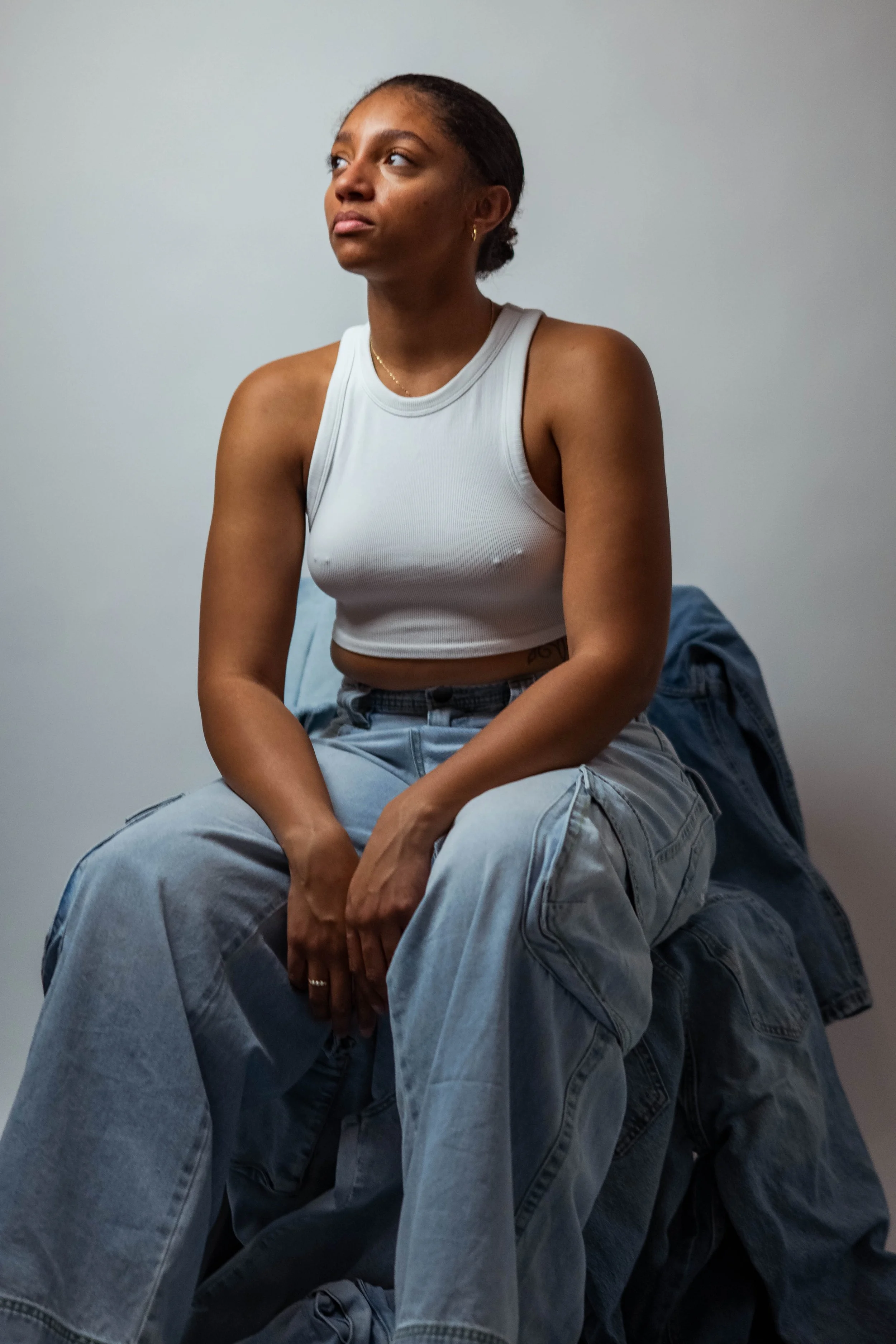 A young woman with brown skin and dark hair tied back, wearing a white sleeveless crop top and baggy light-wash jeans, sitting on a pile of denim jackets against a plain gray wall.