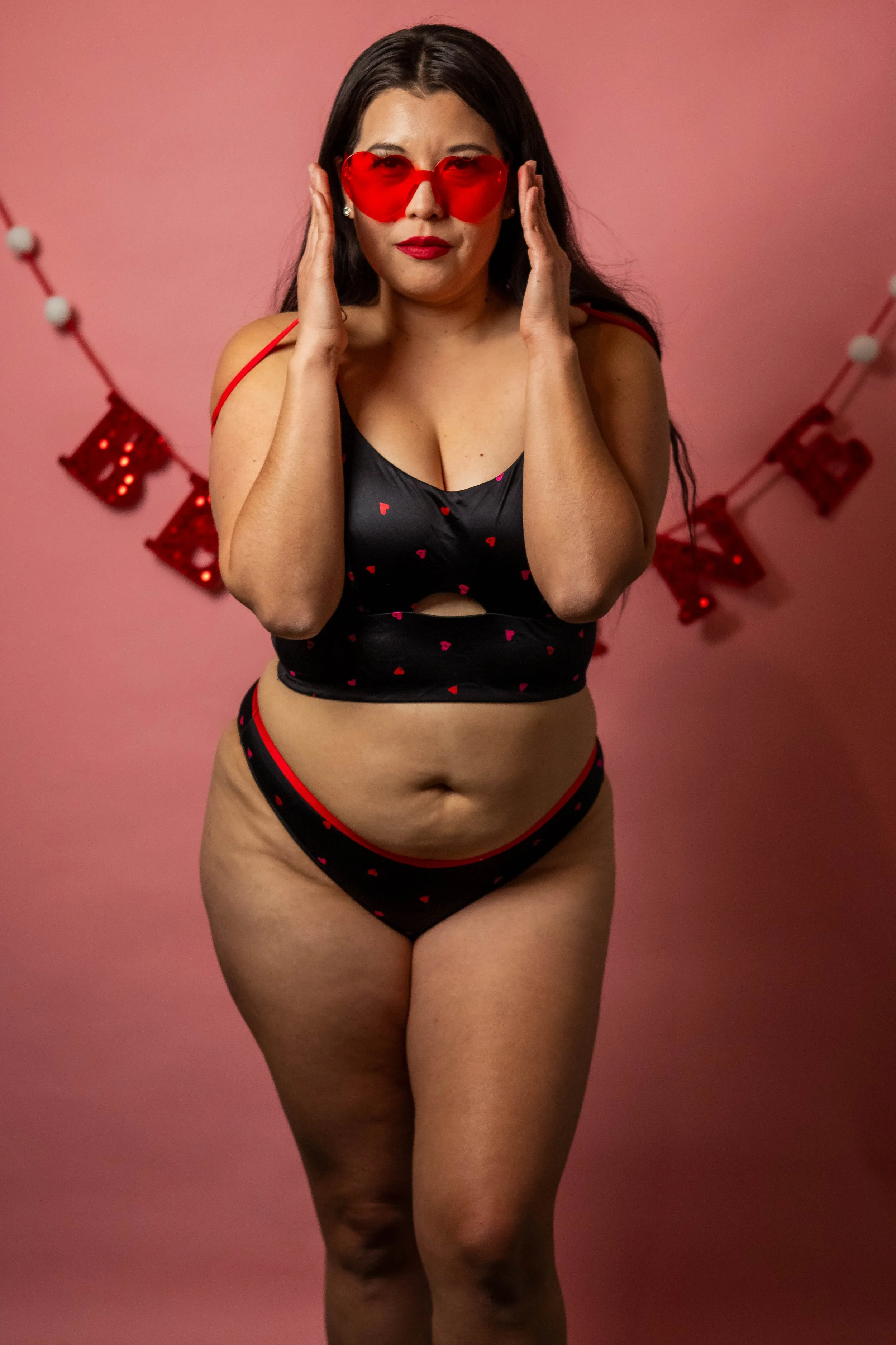 A woman wearing red heart-shaped sunglasses, black lingerie with red hearts, and posing in front of a pink wall decorated with red Valentine's Day ornaments.