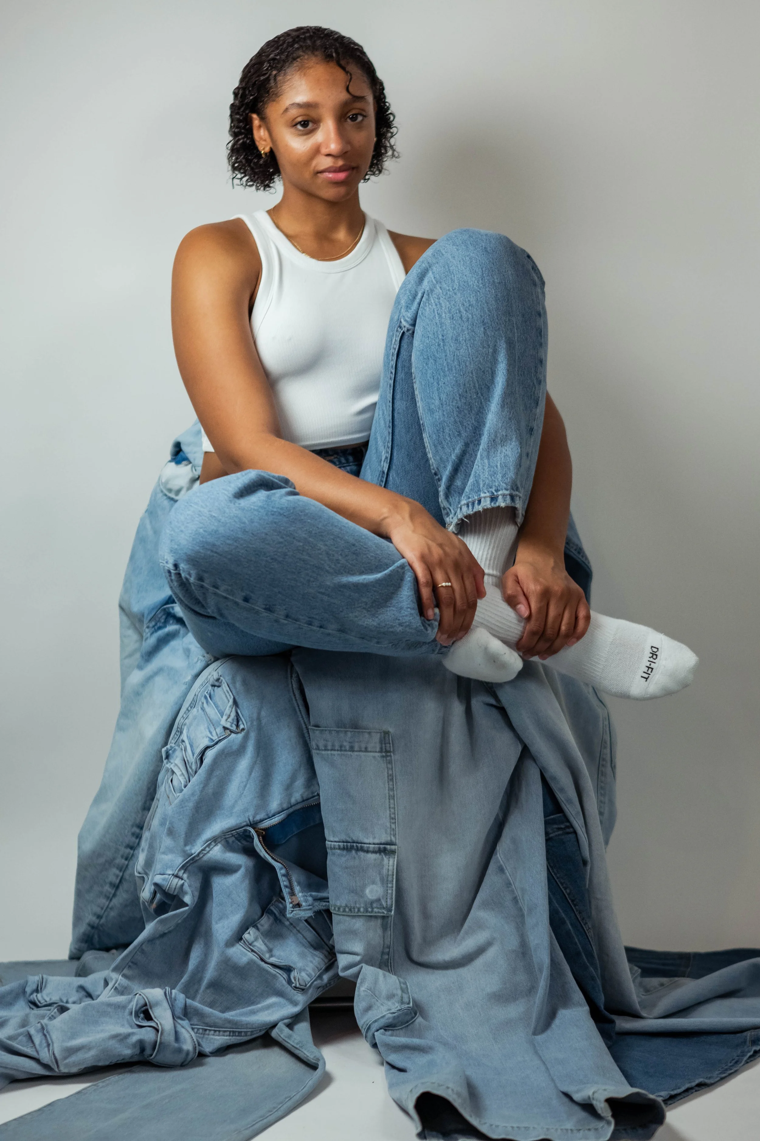 A woman with curly hair, wearing a white sleeveless top, blue jeans, and white socks, sits on a pile of denim jeans against a plain white wall.