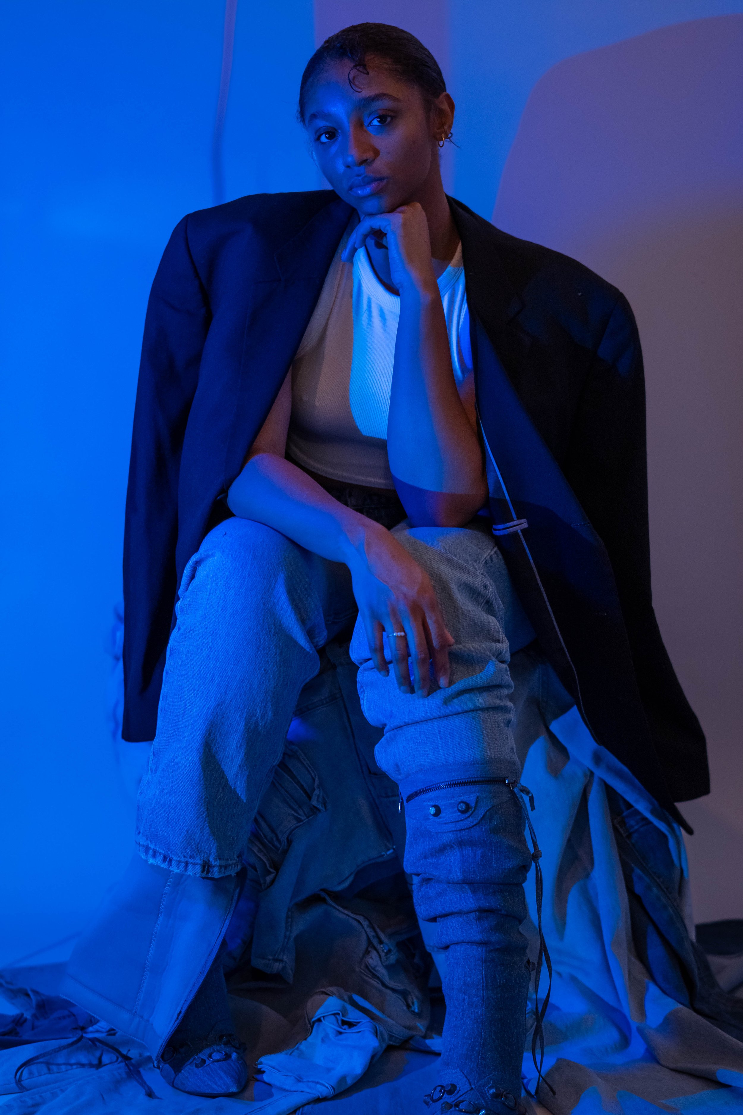 A young woman with short hair sits in a dimly lit room illuminated by blue light. She wears a blazer draped over her shoulders, a white top, and loose jeans, with one knee raised and resting her chin on her hand. There is a pile of clothes and shoes 