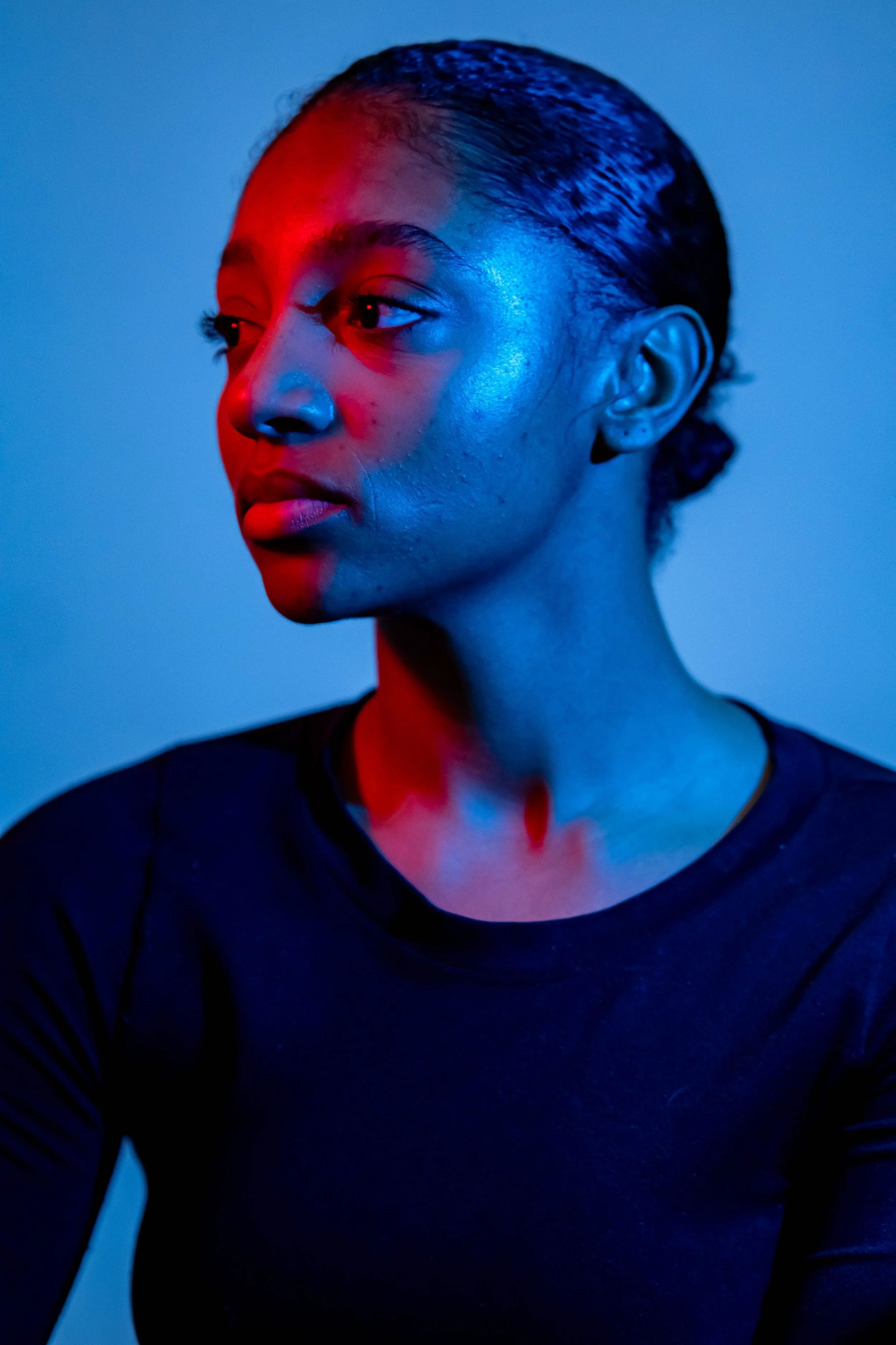 A woman with dark hair slicked back, illuminated by red and blue lighting, looking to the side with a serious expression, wearing a black top.
