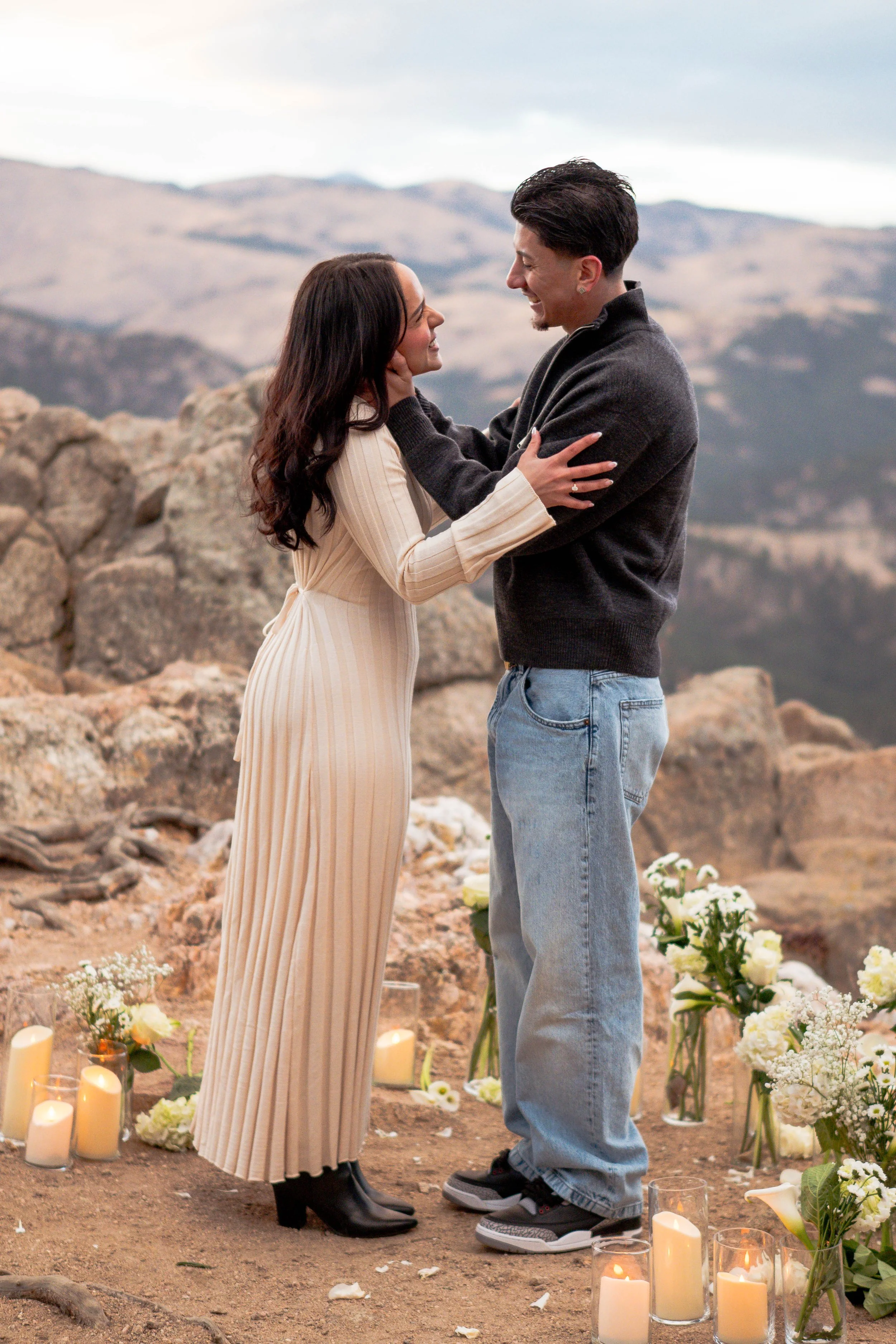 A couple is standing close together during a wedding ceremony outdoors with mountains in the background, surrounded by candles and white flowers.
