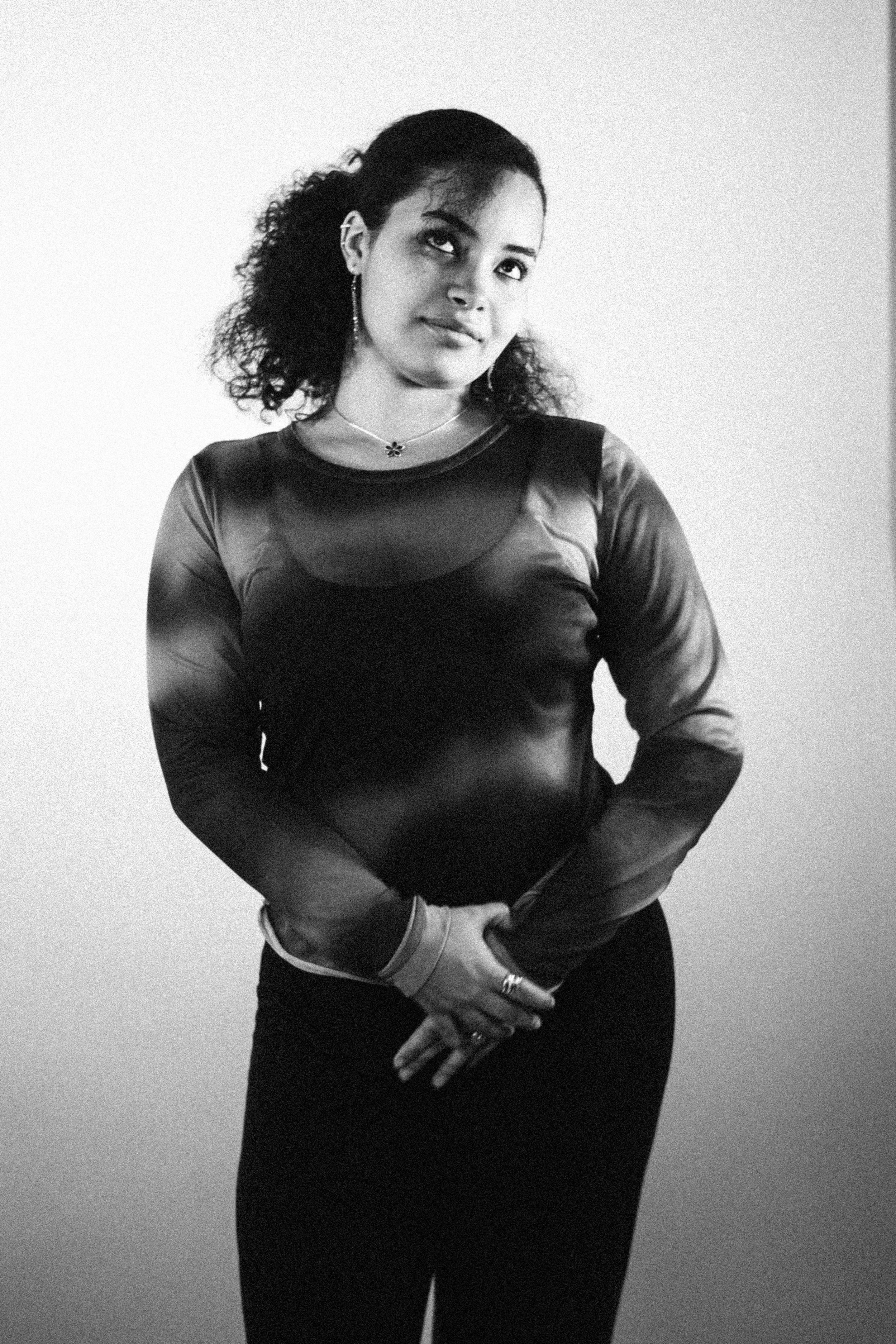A woman in a long-sleeved sheer top with a dark skirt, standing with hands clasped in front, against a plain background.
