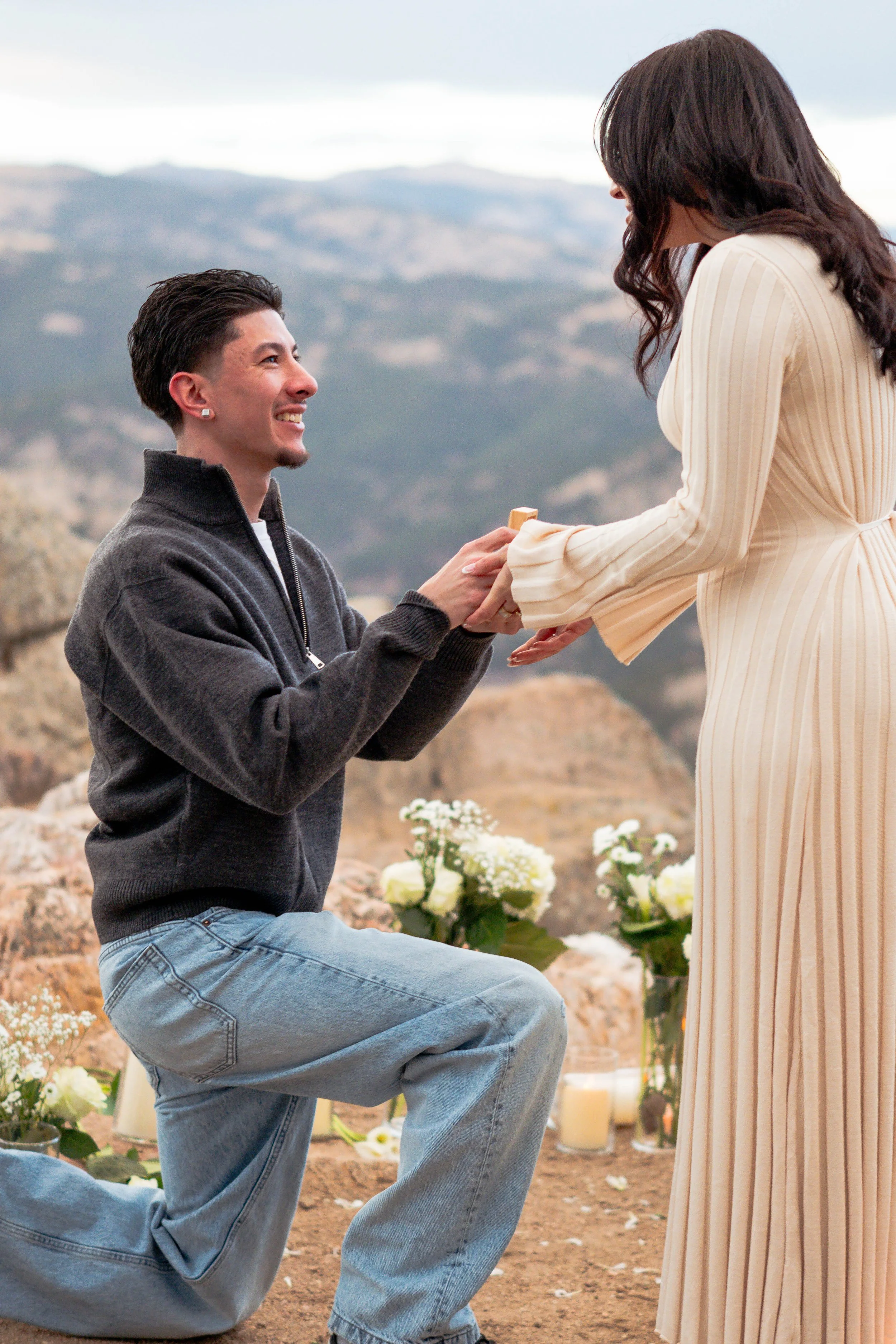 A man proposing marriage to a woman outdoors with mountains in the background, surrounded by flowers and candles.
