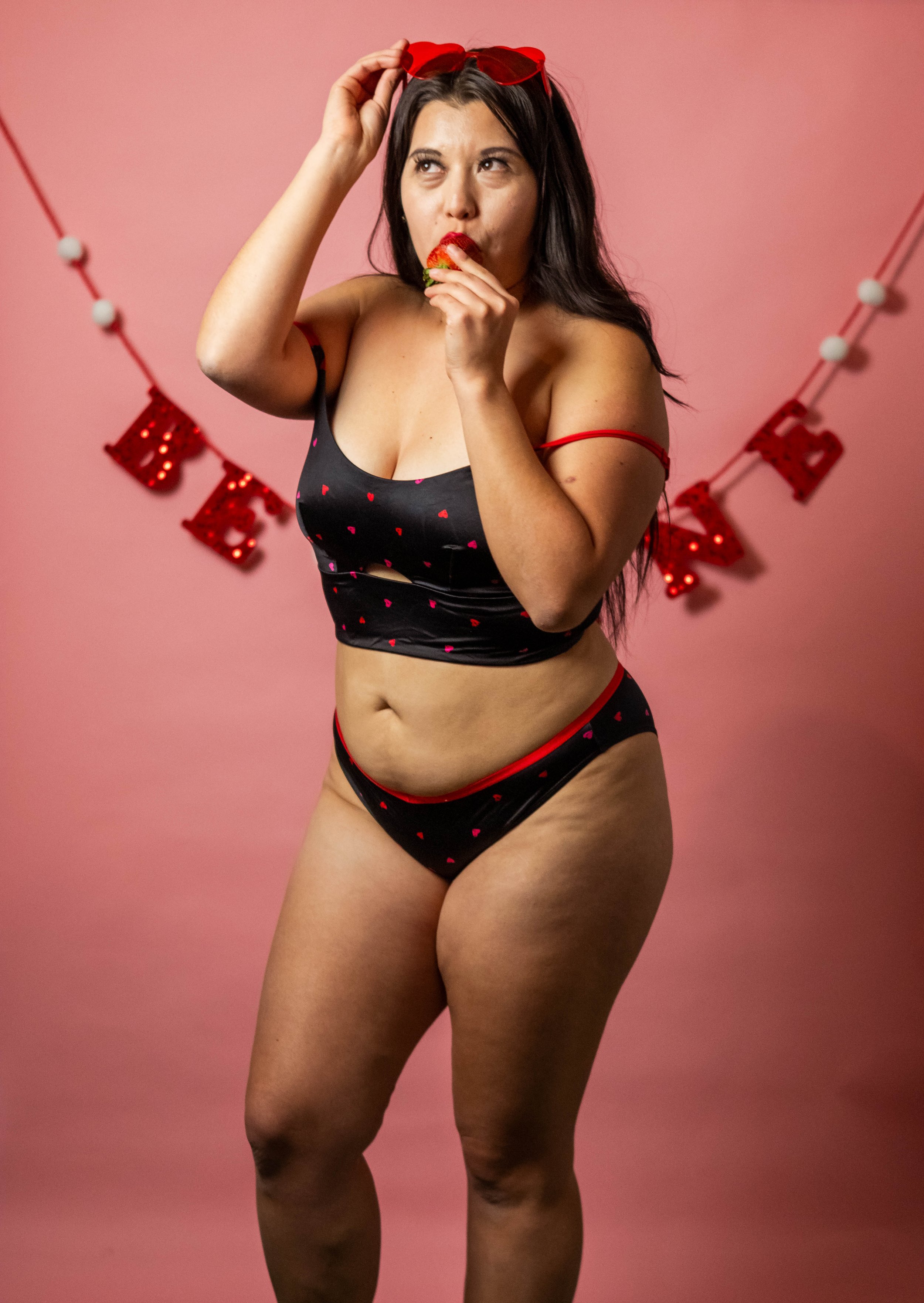 Woman in black lingerie with red hearts, holding a strawberry near her mouth, standing in front of a pink background with 'LOVE' decoration.