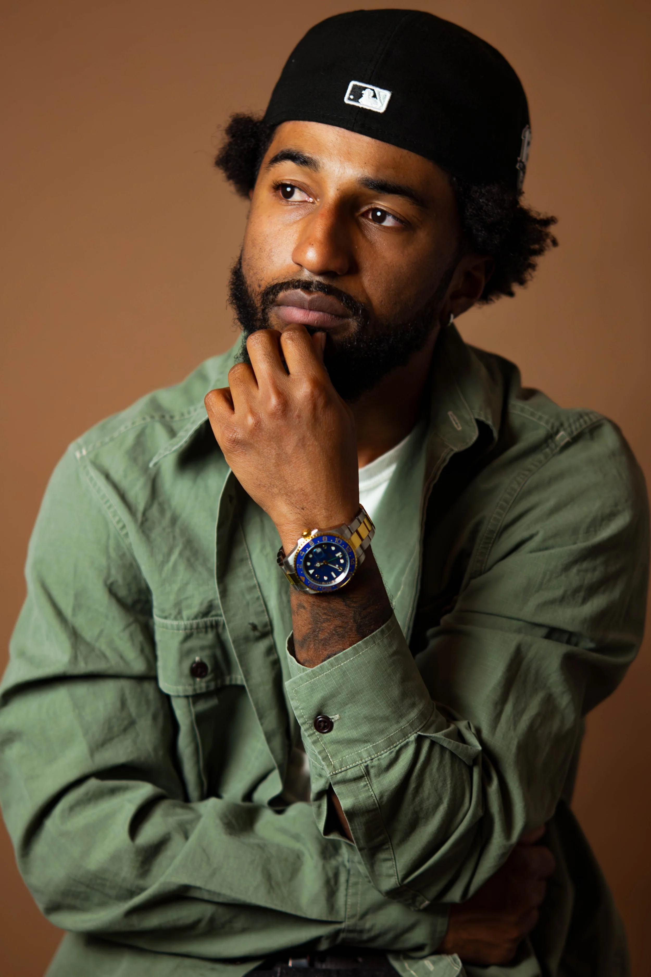 A man in a black baseball cap, green shirt with rolled sleeves, and a gold and blue watch, thinking with his hand on his chin against a plain brown background.