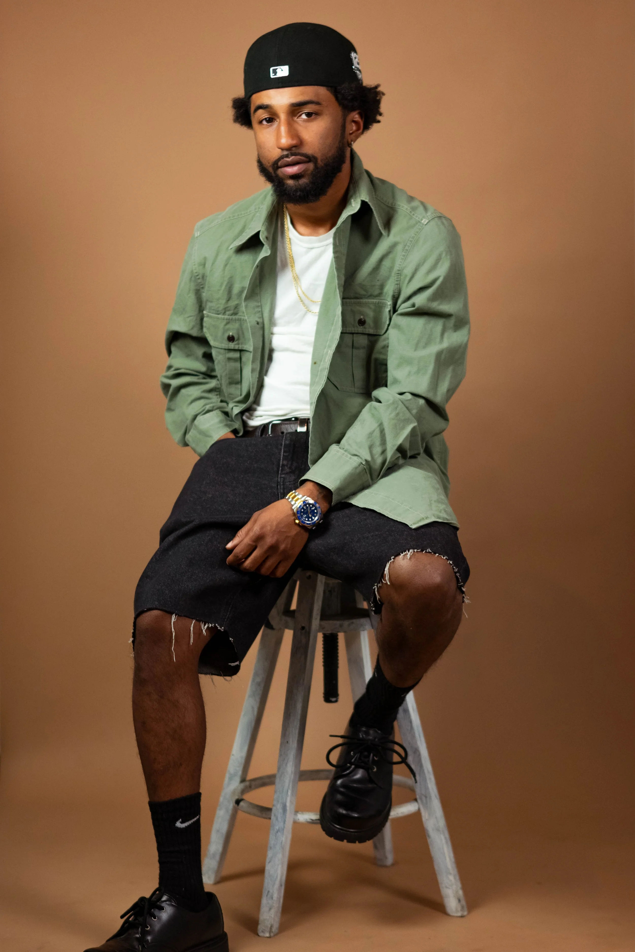A young man with a beard and long curly hair sitting on a wooden stool against a plain brown background. He is wearing a black baseball cap backwards, a green jacket over a white T-shirt, ripped black shorts, black sneakers, and black socks. He has a