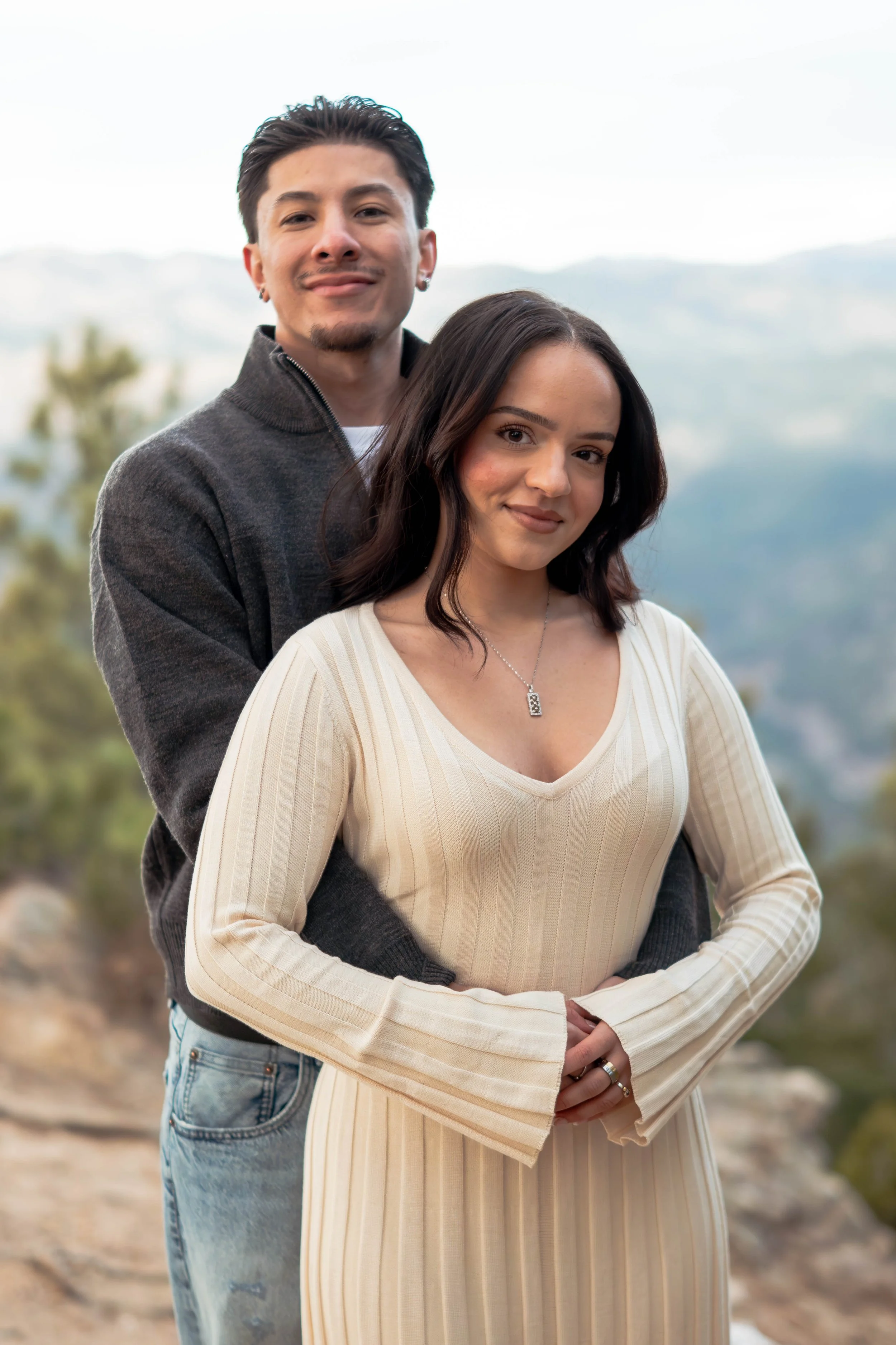 A young couple outdoors, with mountains and trees in the background, standing close together. The man is behind the woman, with his arms around her waist, both smiling softly at the camera.