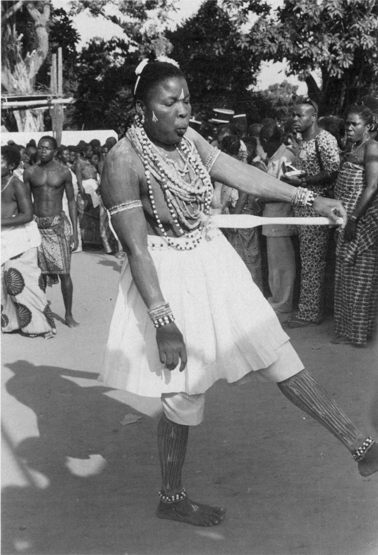   A Mami Wata medium, a trident paddle on her hip, lines, and tridents painted on her body with powdered white clay, and rainbow serpent beads hung around her neck-leans in an off-balance pose during trance before beginning her dance. Togo, Mina, 197