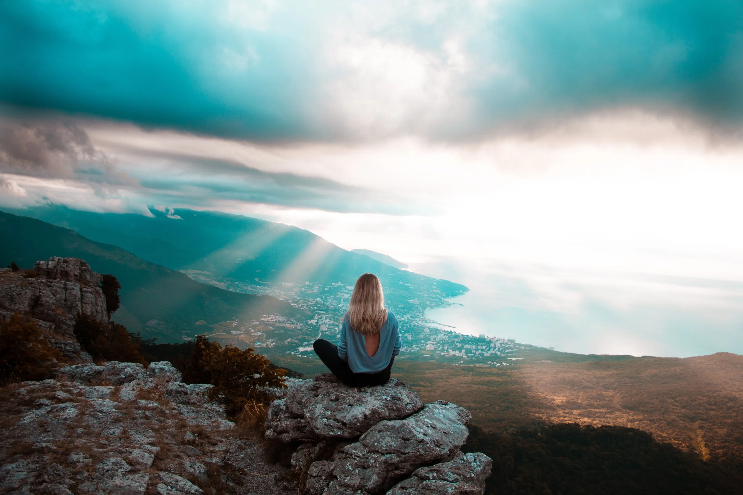 A woman sitting on rocks on a mountain, overlooking a valley, with dramatic clouds and sunlight breaking through