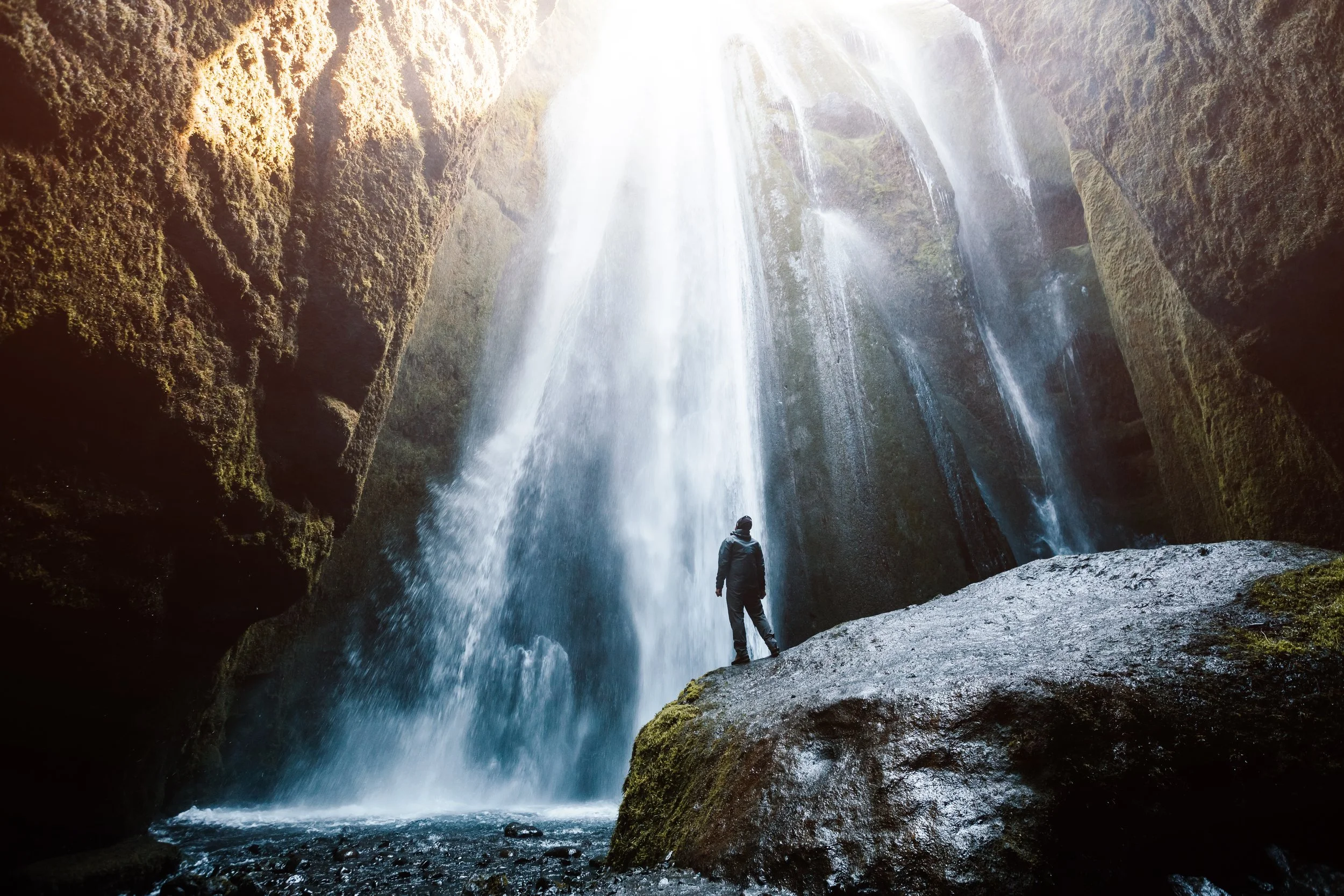 A person standing on a rock in front of a waterfall inside a cave with sunlight shining through.