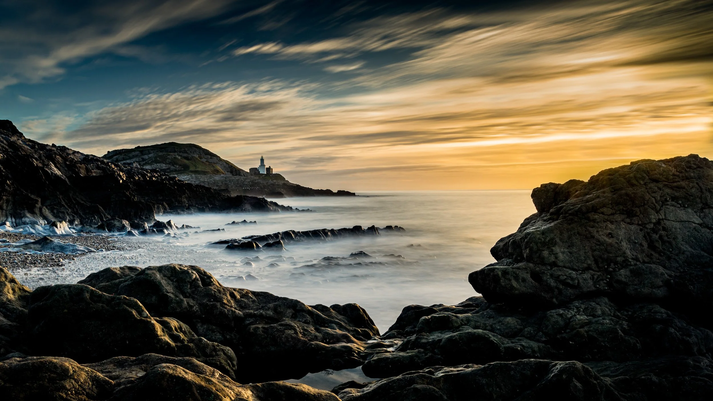 Sunset over a rocky coastline with a lighthouse on a distant hill