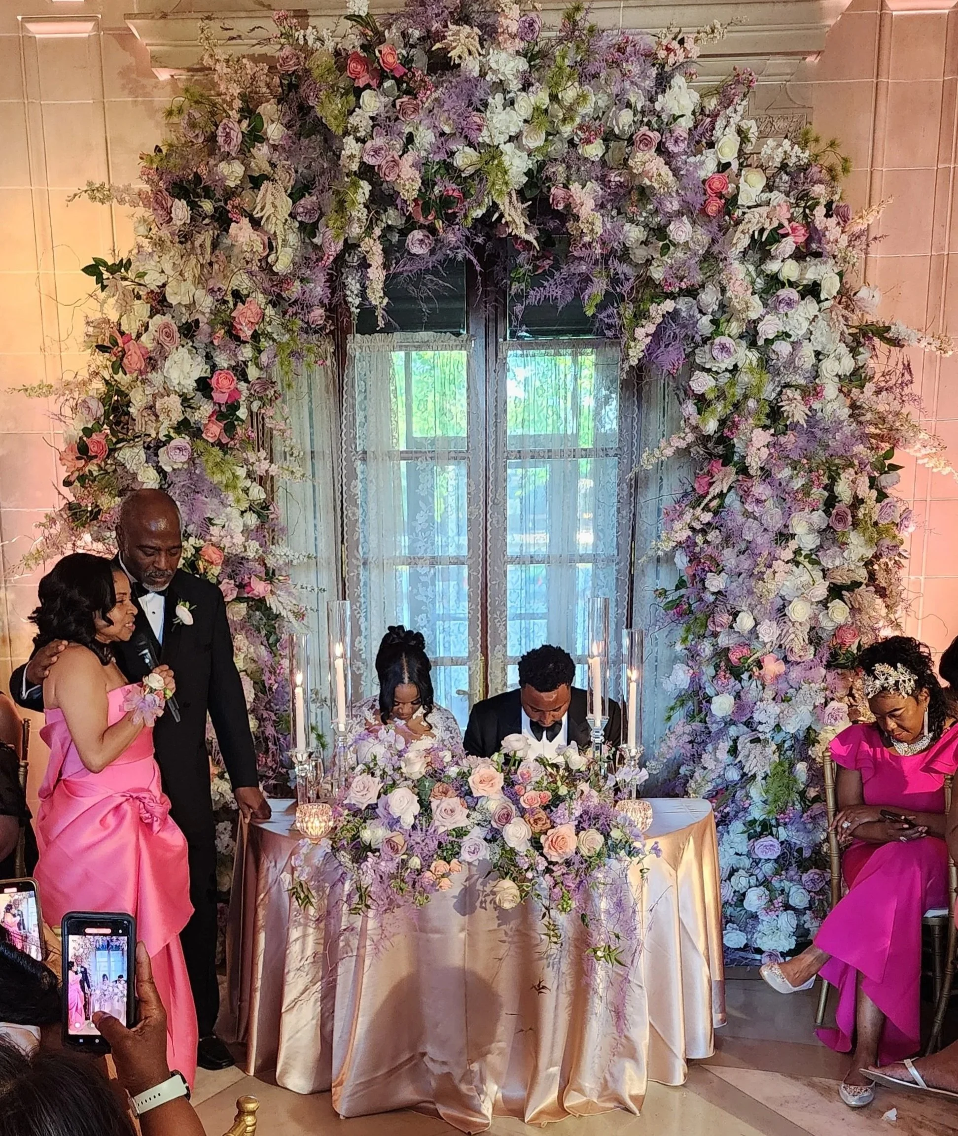 A wedding ceremony with a floral arch, a bride and groom signing documents, and guests dressed in pink and black.