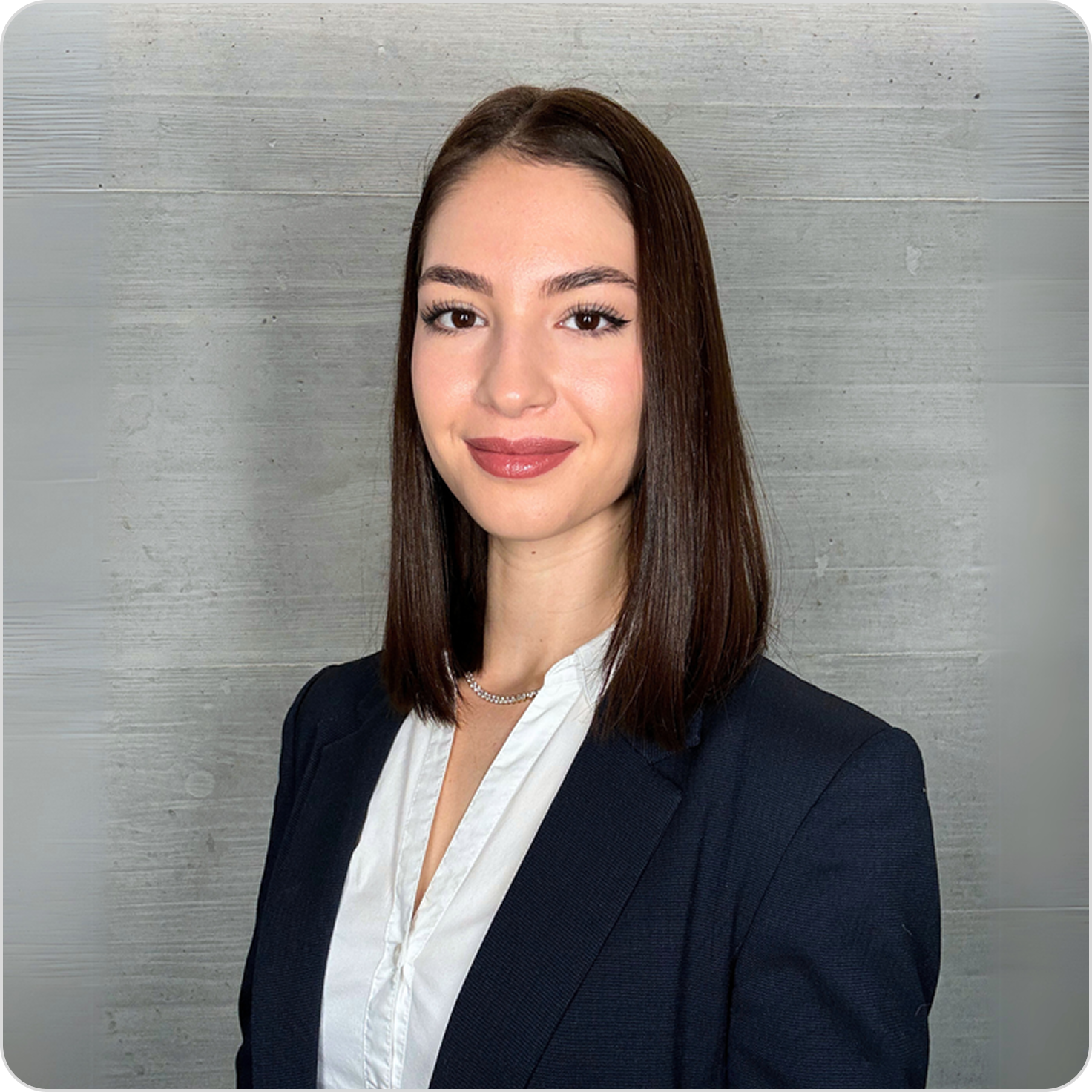 Professionelles Porträt einer jungen Frau mit braunen Haaren, dunklem Anzug und weißer Bluse vor einer grauen Holzwand.