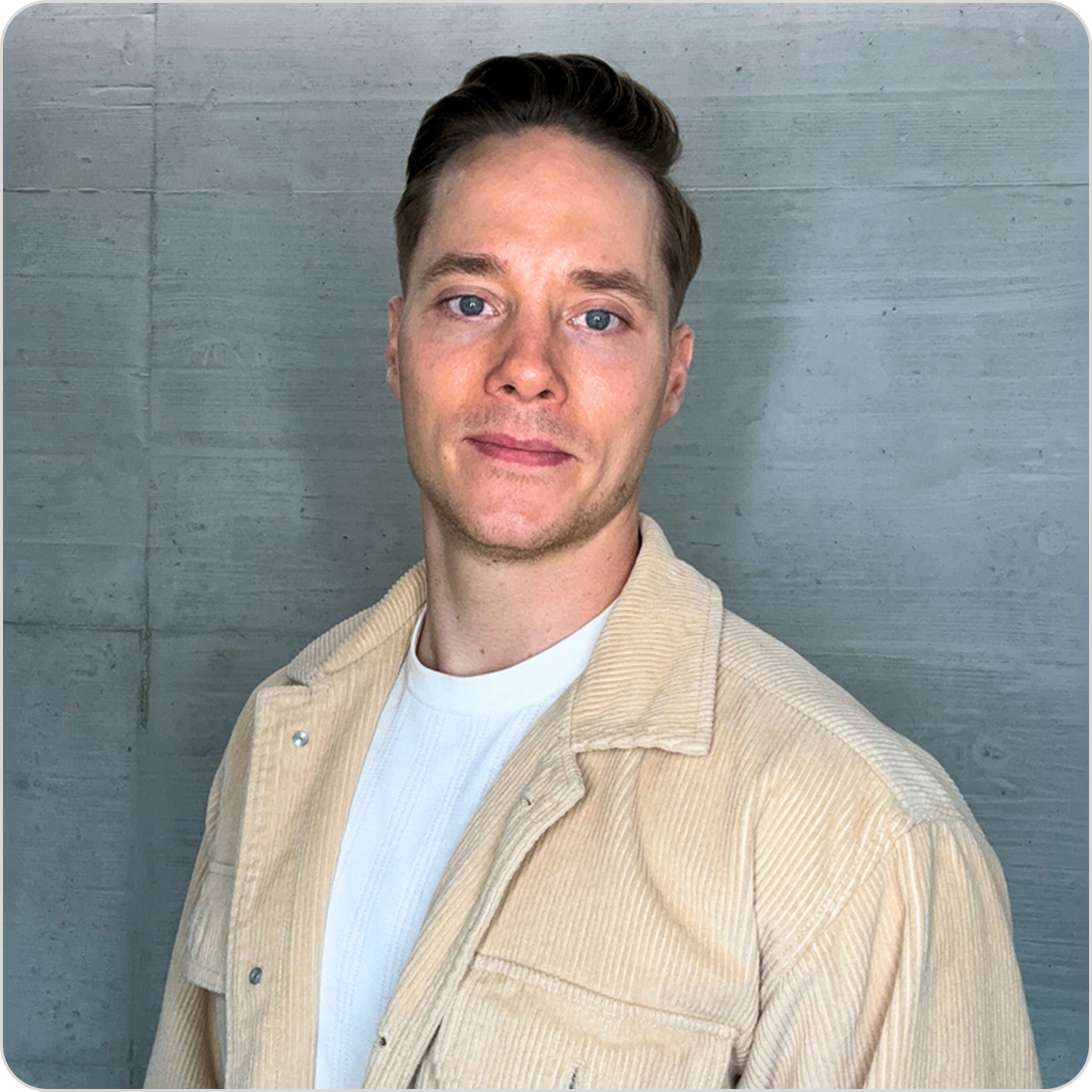 Ein junger Mann mit kurzem braunem Haar, blauen Augen, heller Haut und einem leichten Lächeln steht vor einer grauen Betonwand.