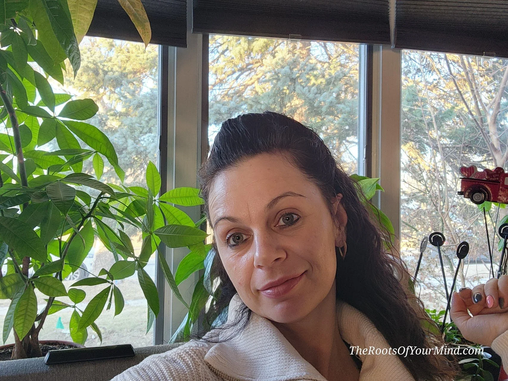 A woman with dark curly hair and light skin, wearing earrings and a cream-colored top, posing indoors near large windows and green plants.