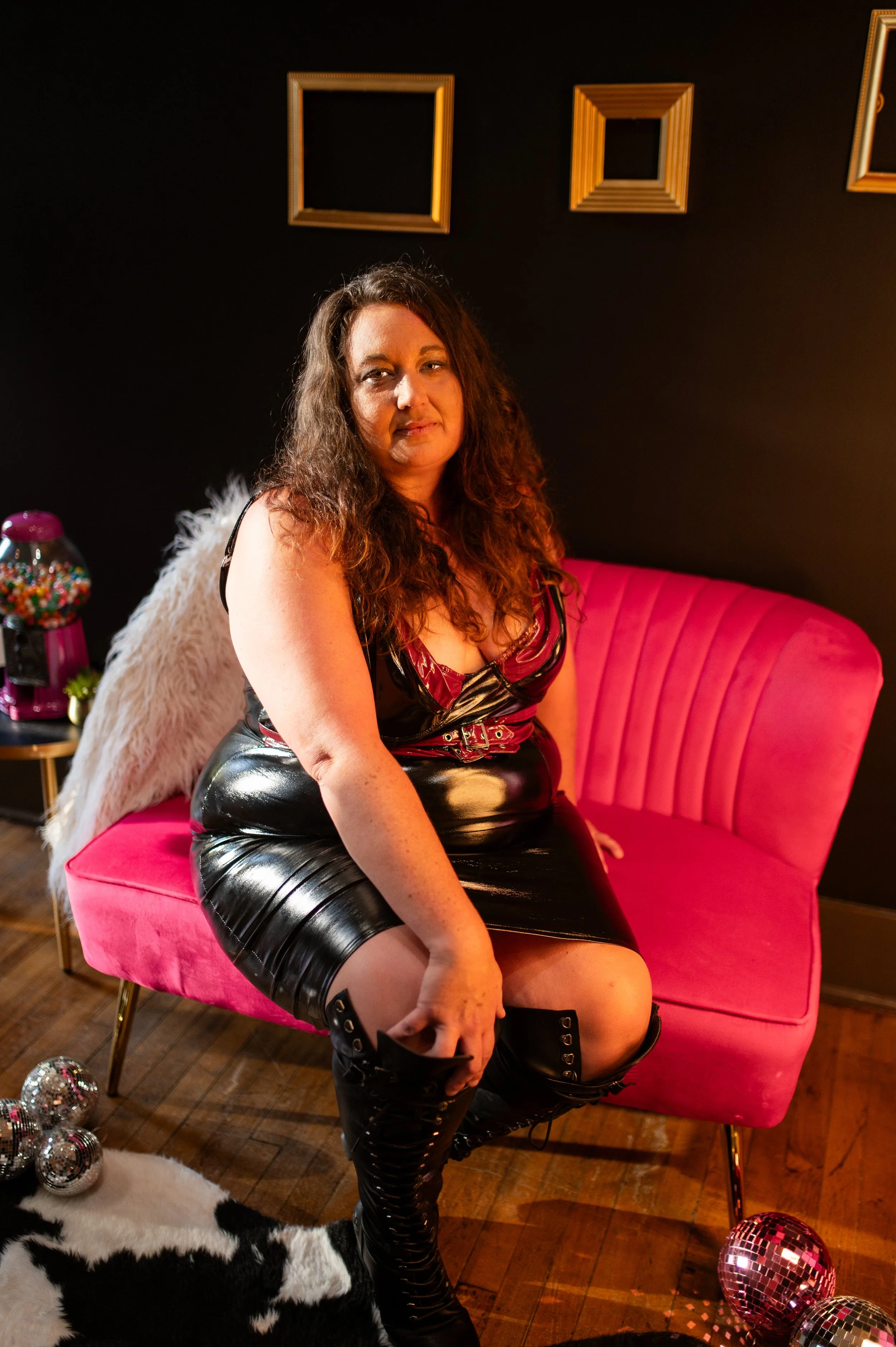 A woman with long curly hair wearing a black and red vinyl dress, with angel wings, sitting on a pink velvet sofa in a room decorated with disco balls and framed art on a black wall.