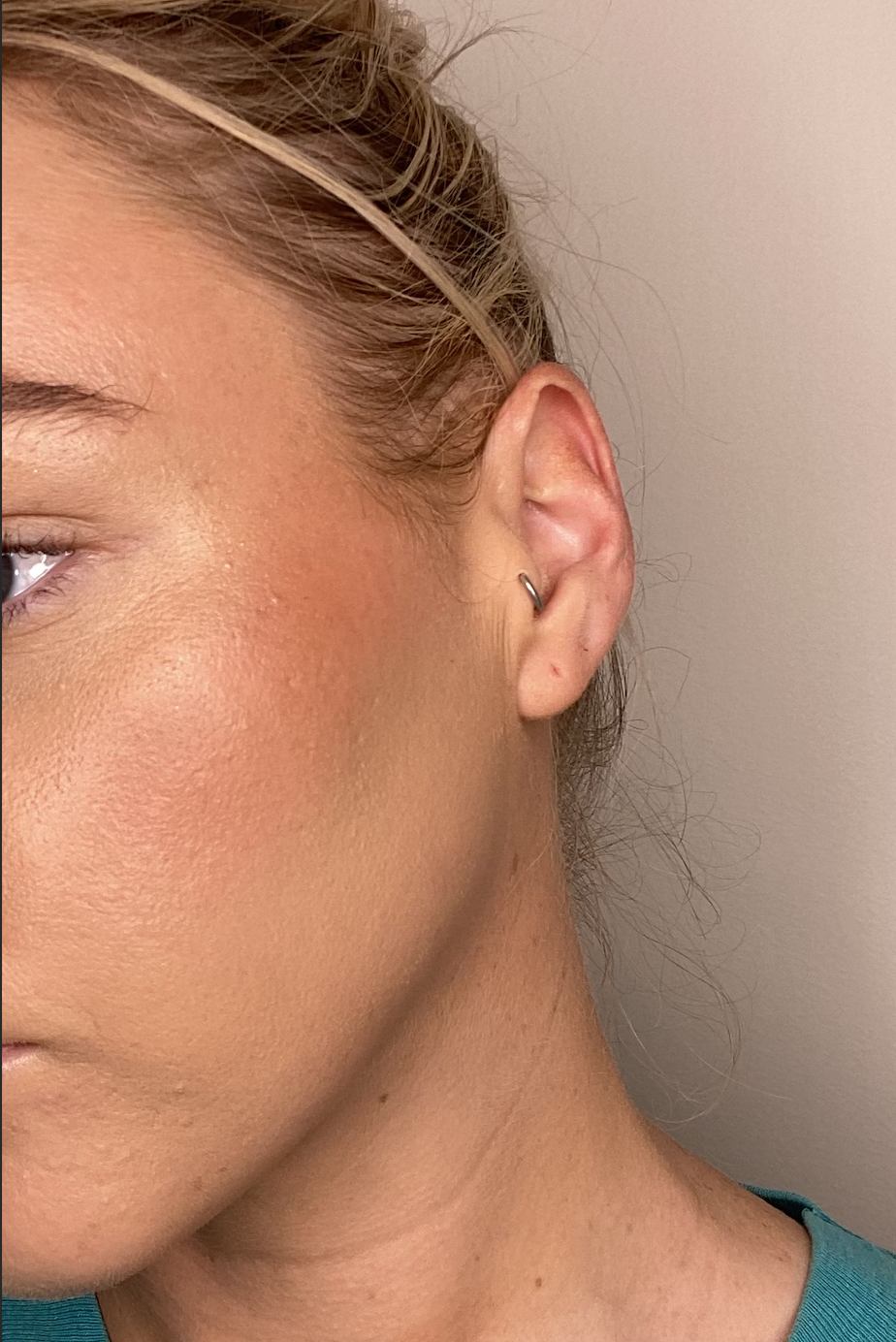 Close-up of a woman's right side of face showing her ear with a small silver hoop earring, part of her eyebrow, cheek, and neck, with her hair pulled back.