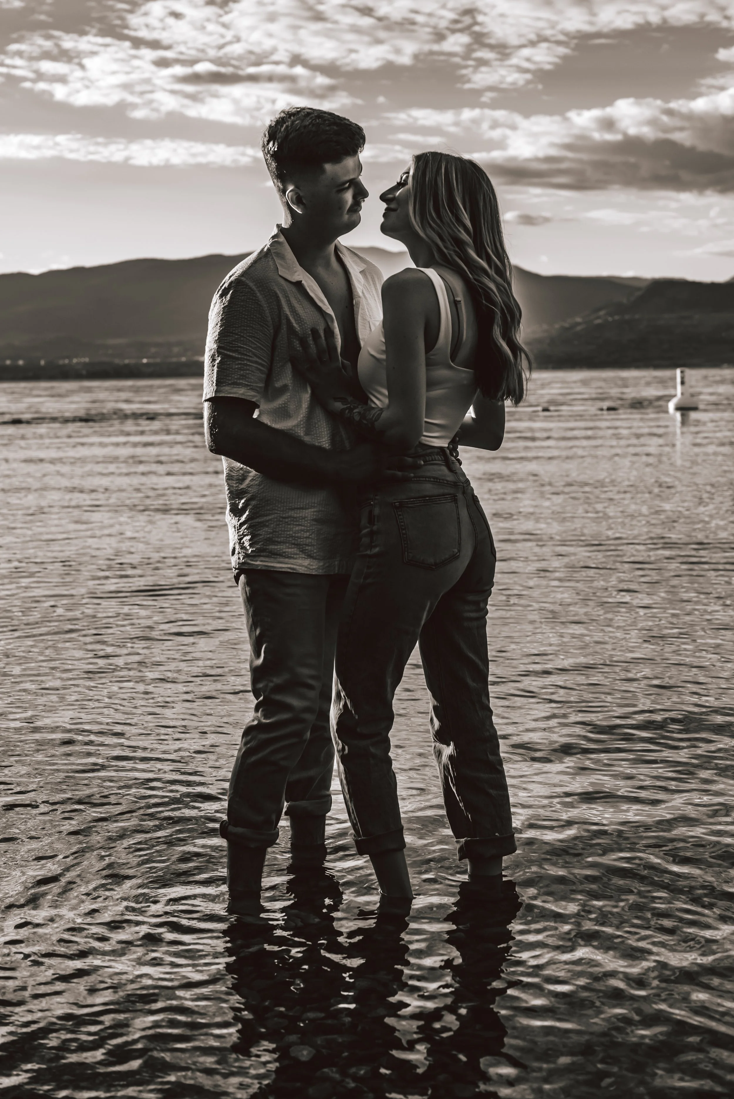 A black-and-white photo of a young couple standing in shallow water at a lake or river, gazing into each other's eyes, with mountains and a cloudy sky in the background.