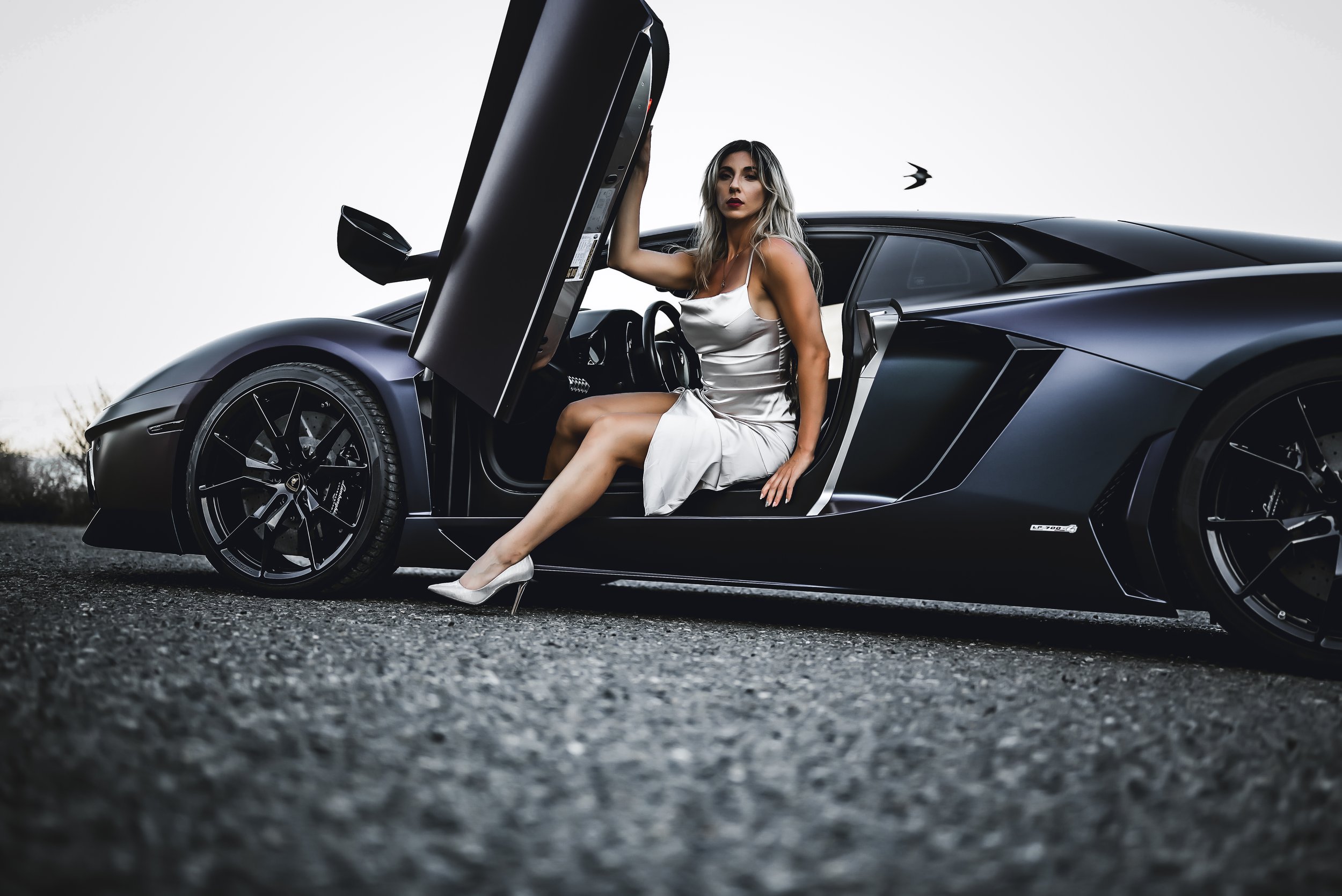 A woman in a satin dress and high heels sitting in a black luxury sports car with the door open, outdoors on a paved surface.