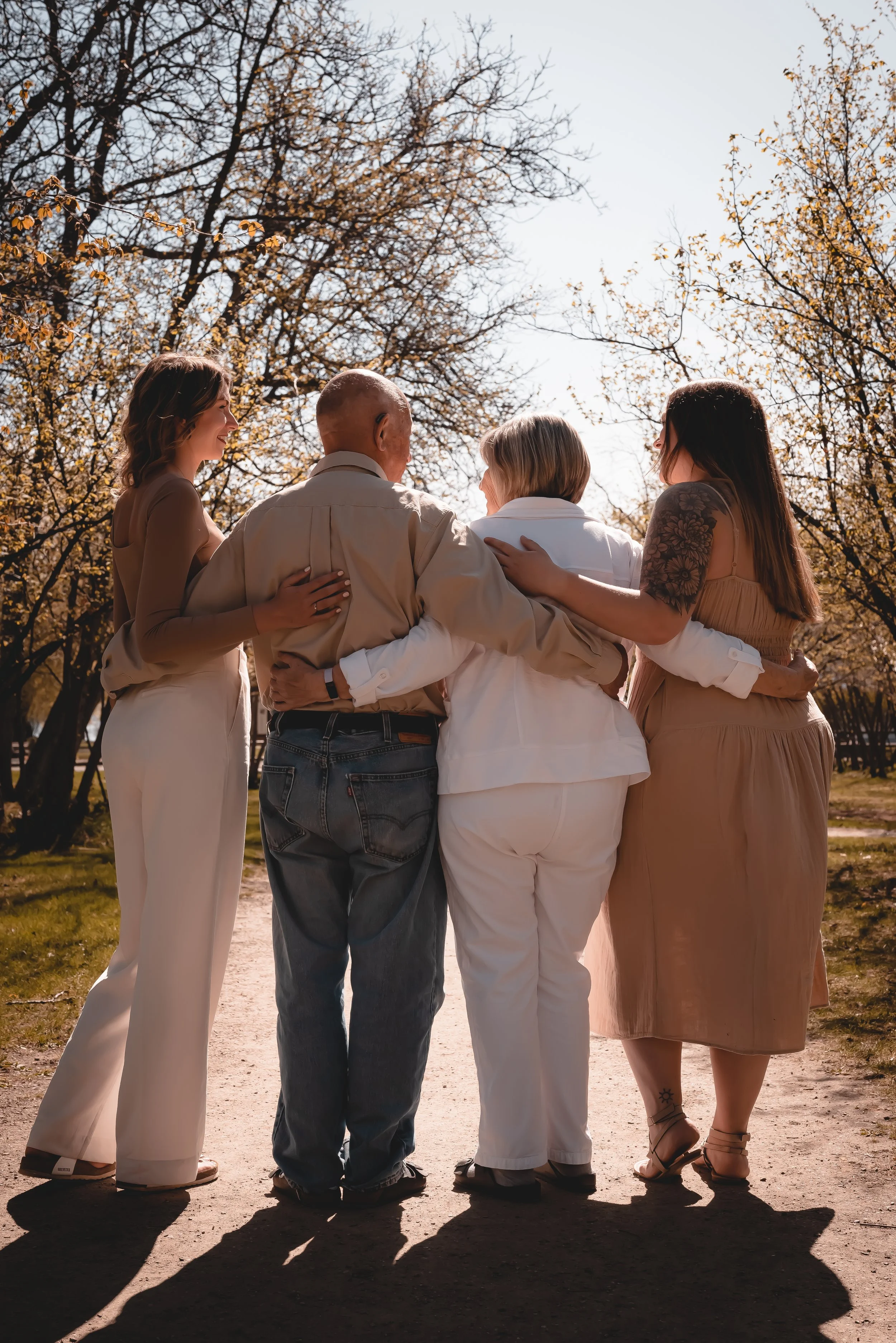 Four women and one man standing outdoors on a sunny day, embracing and walking together on a dirt path surrounded by trees with budding leaves.