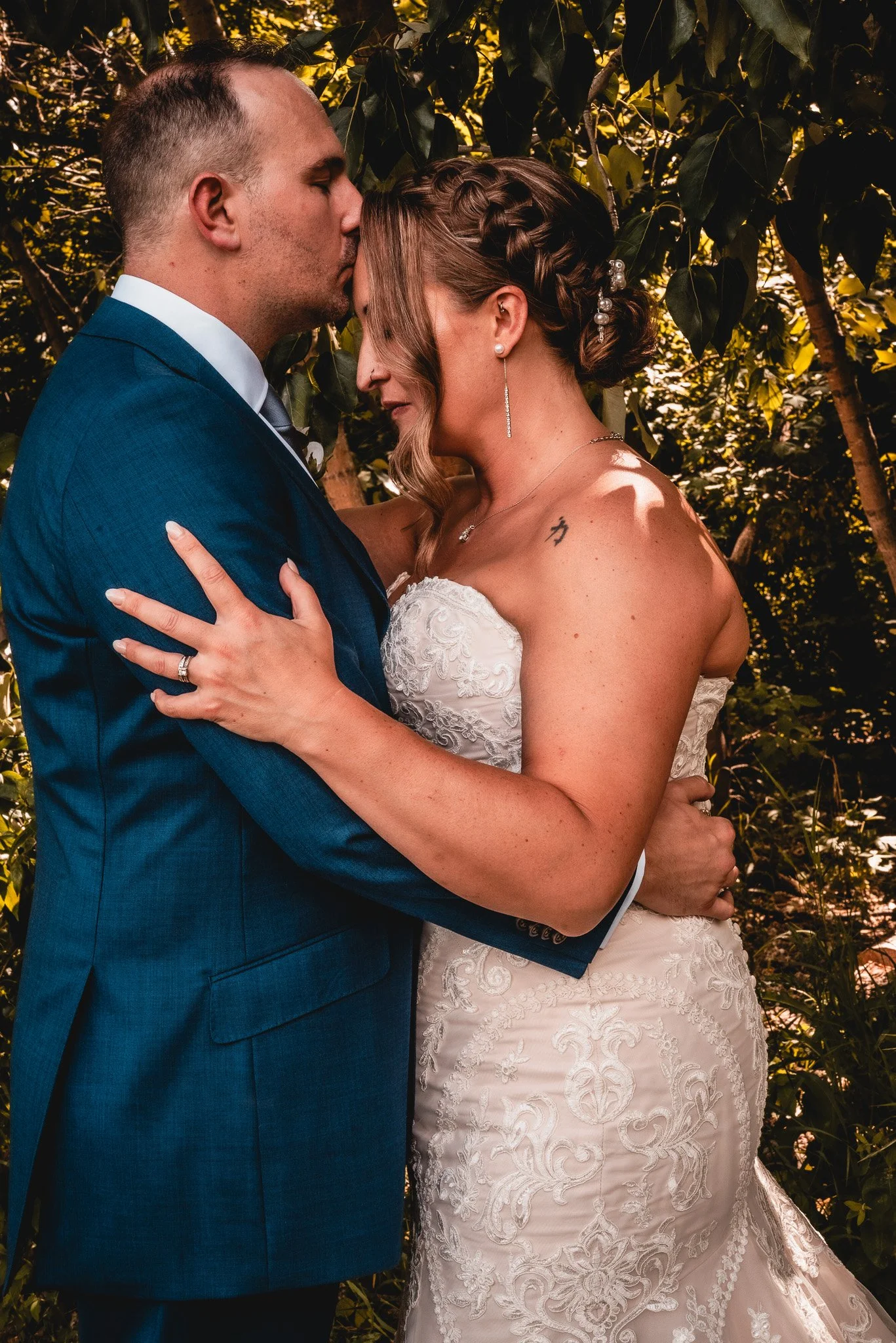 A bride and groom embrace and share a kiss outdoors among green leaves, with the groom in a blue suit and the bride in a white lace wedding dress.