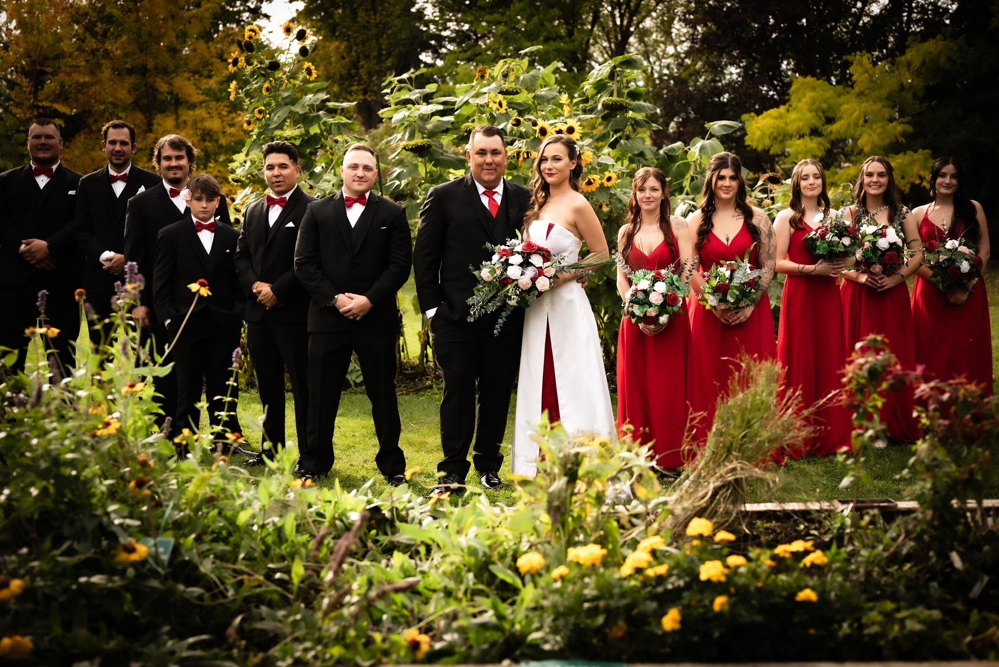 A wedding party stands outdoors in front of sunflowers and trees, with the bride and groom at the center. The bride wears a white gown holding a bouquet, and the groom wears a black suit with a red tie. The bridesmaids in red dresses hold bouquets, and the groomsmen in black suits with red bow ties stand on either side.