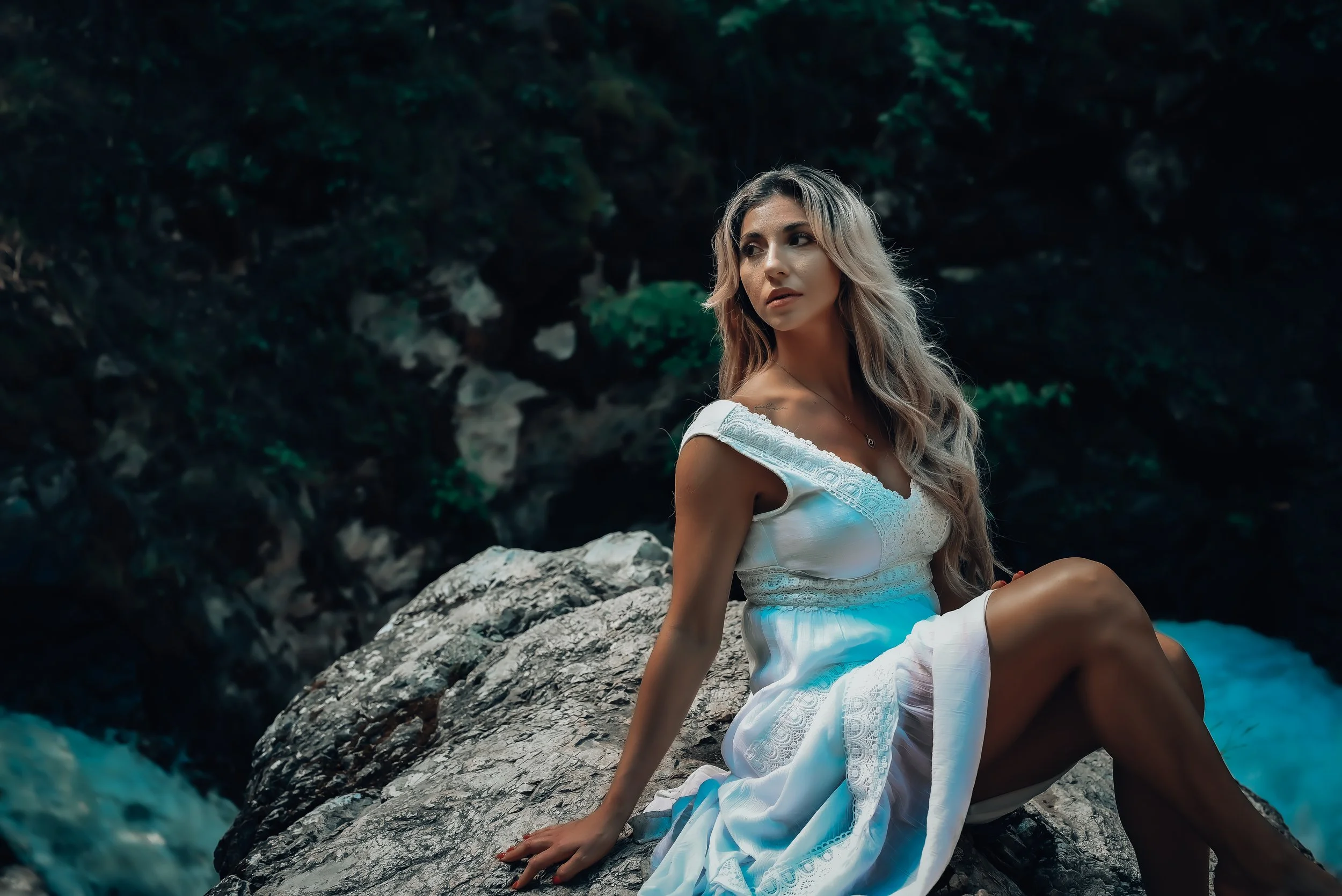 A woman with long blonde hair sitting on a large rock near a waterfall in a forest, wearing a white dress.