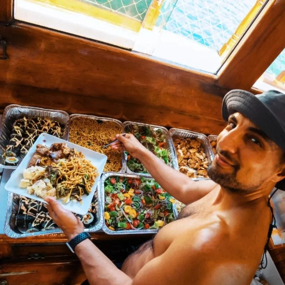 A guest on a traditional wooden junk boat holding a plate of food from a diverse catering buffet featuring noodles, salads, and skewers