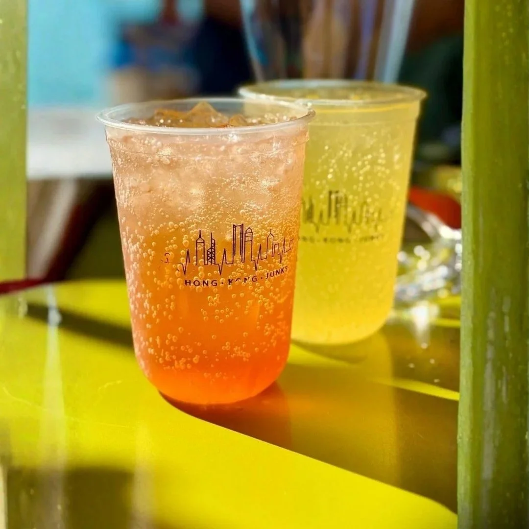 Two refreshing signature fruit sodas in branded Hong Kong Junks cups, showing vibrant orange and yellow colors with condensation on a sunny boat deck.
