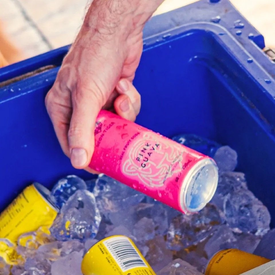A close-up shot of a hand pulling a bright pink Neon Tiger Pink Guava real fruit soda can out of a blue ice-filled cooler.
