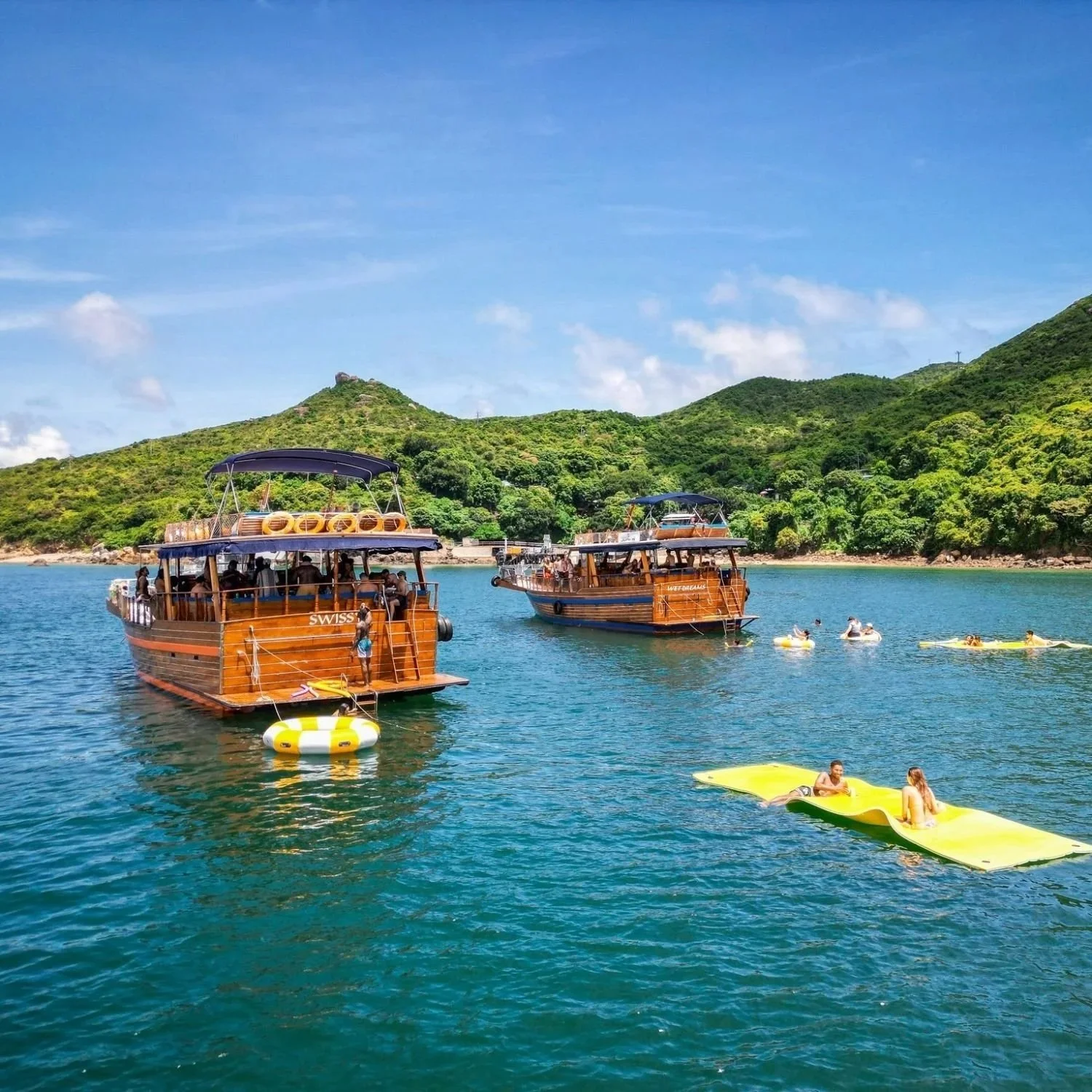 Two traditional teak wood junk boats anchored together for a large group event, with guests enjoying the water and a yellow floating mat in a scenic Hong Kong bay.