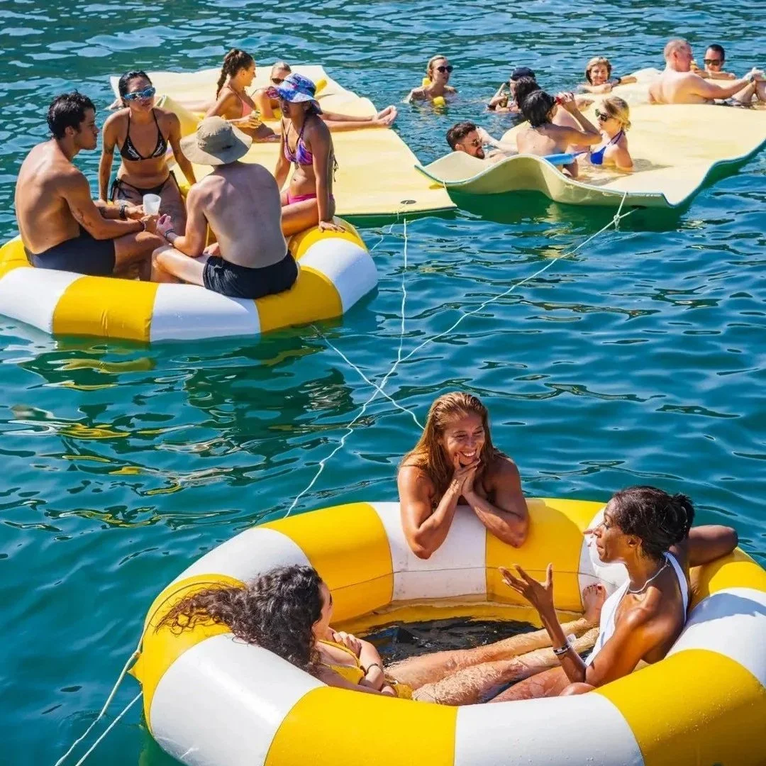Guests relaxing and socializing on large yellow floating mats and circular inflatable tubes in a calm Hong Kong bay.