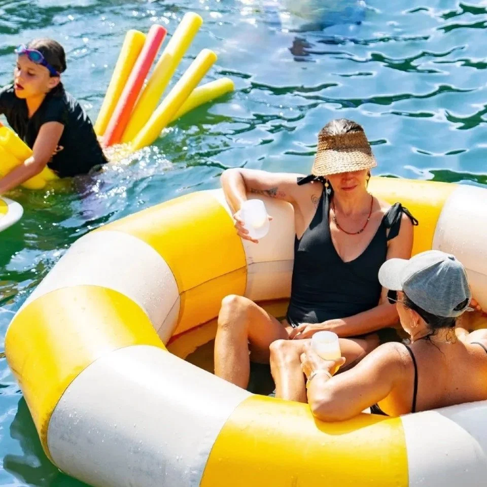 Two women enjoying drinks while sitting in a yellow and white inflatable floating ring in the water.