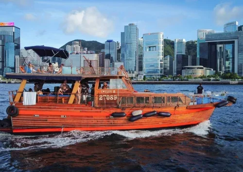 A bright orange boat with people on board, cruising on a river with a city skyline in the background. The boat has the word 'Swissy' and the number 27069 on its side.