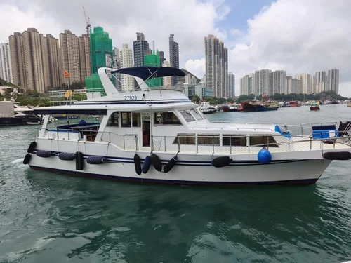 A yacht docked in a marina with a city skyline of tall buildings under a partly cloudy sky in the background.