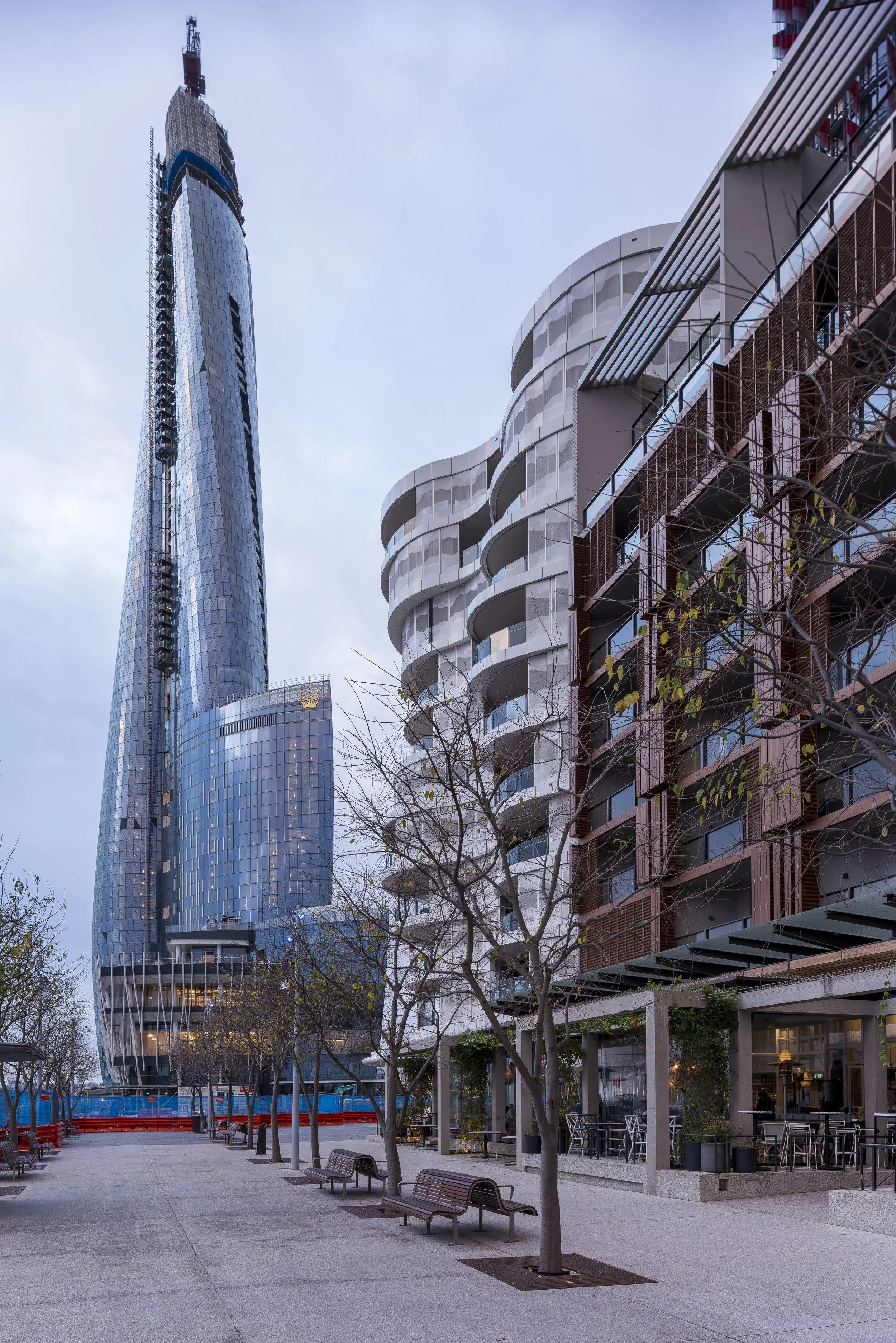 Cityscape with modern skyscrapers, including a tall glass building under construction on the left and a residential building with curved balconies on the right. In the foreground, an empty outdoor plaza with benches, trees, and a few tables.
