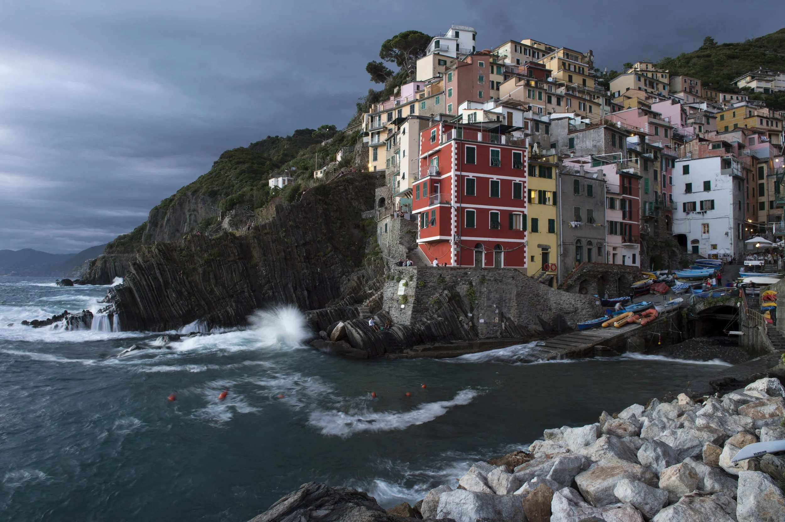 Colorful houses built on a hillside overlooking the ocean, with rocky shoreline and boats docked at a small harbor, under a cloudy sky.