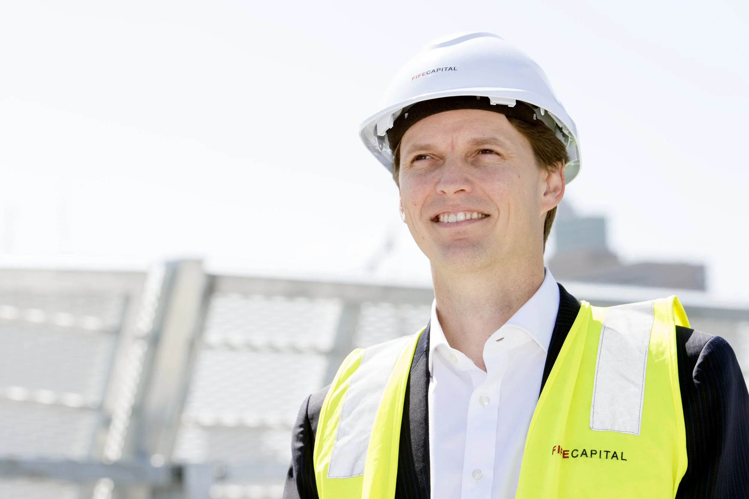 A workman in a white hard hat and yellow safety vest stands outdoors on a construction site, smiling and looking into the distance.