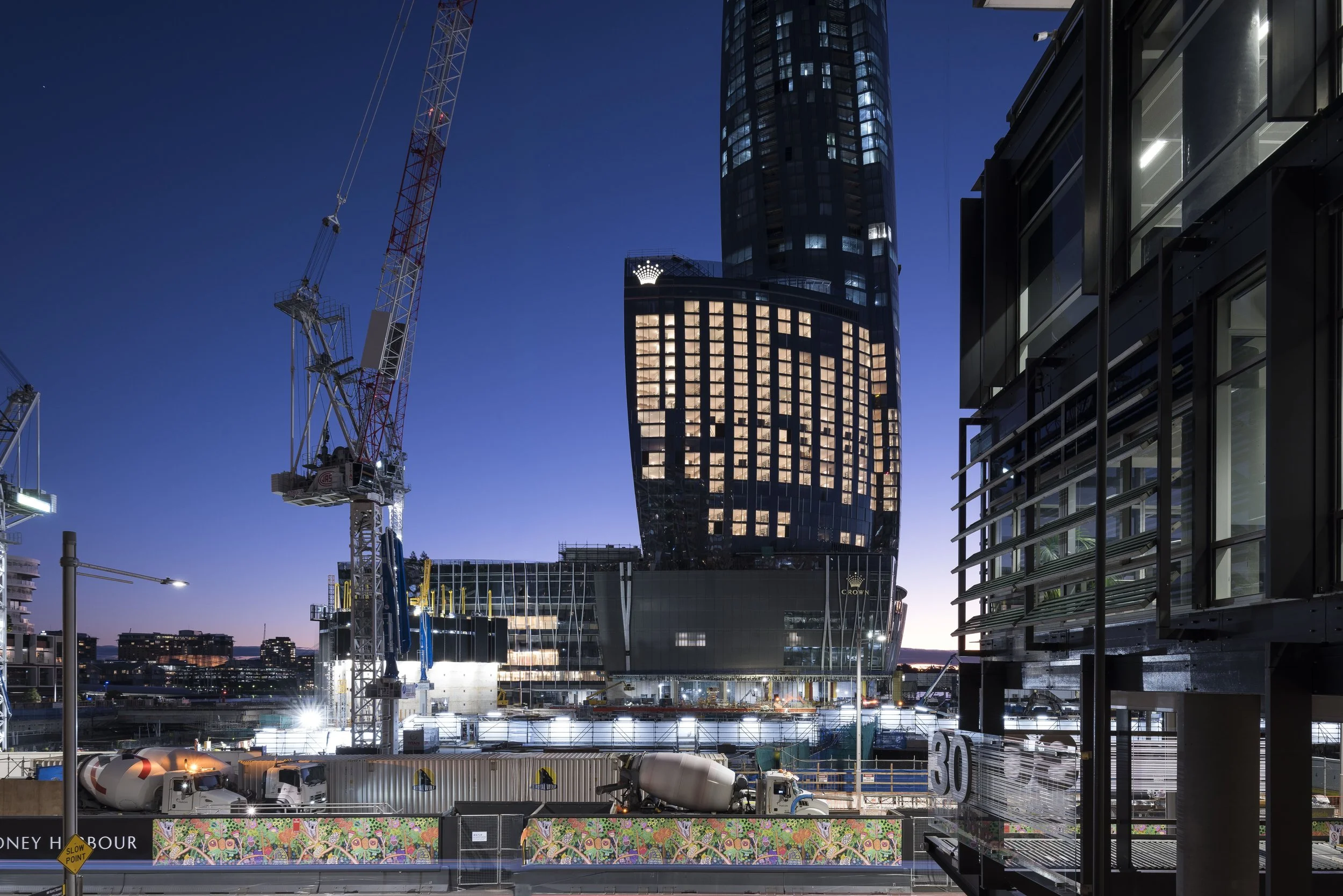 Nighttime view of a construction site in an urban area with tall buildings, cranes, and construction vehicles; illuminated windows in a high-rise building; a colorful mural at the bottom edge of the image; street signs and streetlights in the foreground.