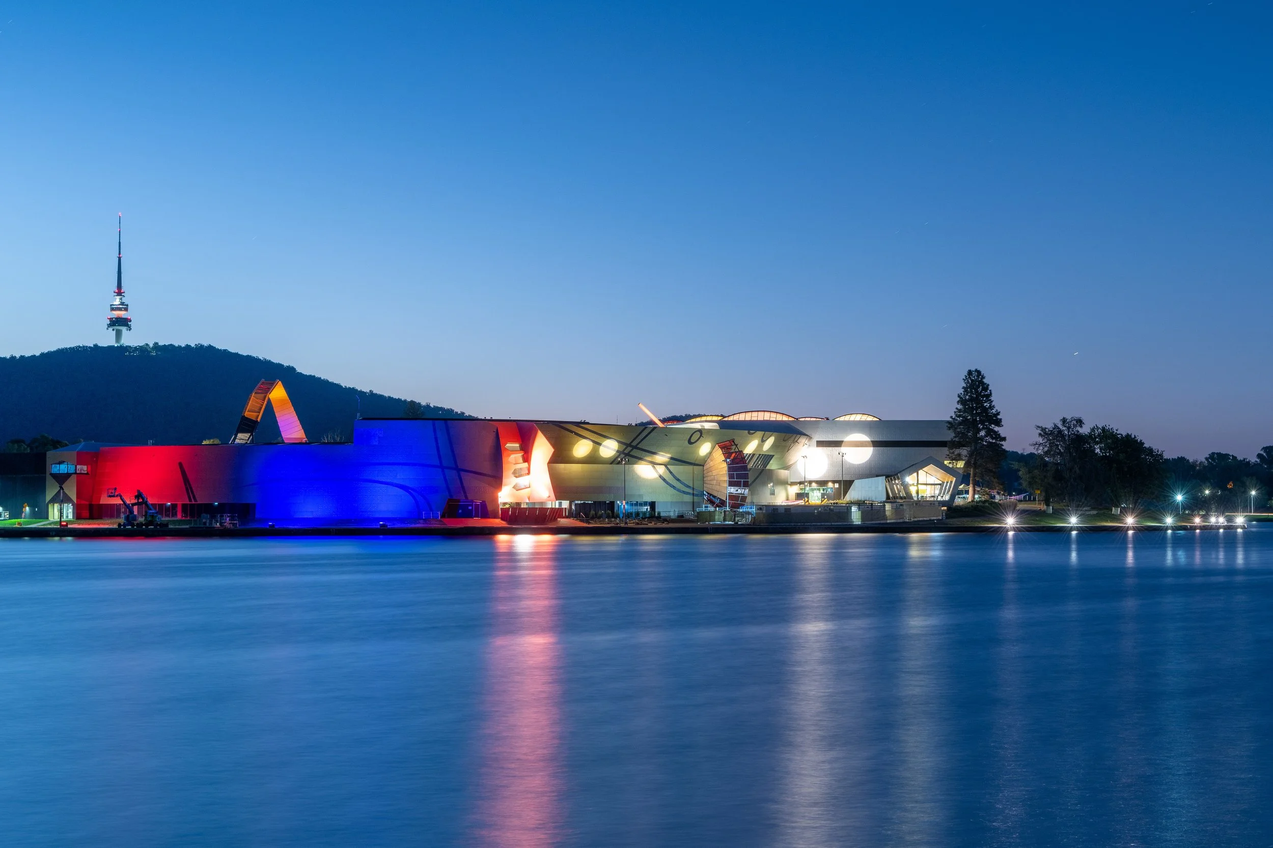 Nighttime view of a large, colorful building on the water with reflections, a hill with a TV tower in the background, and a clear sky.