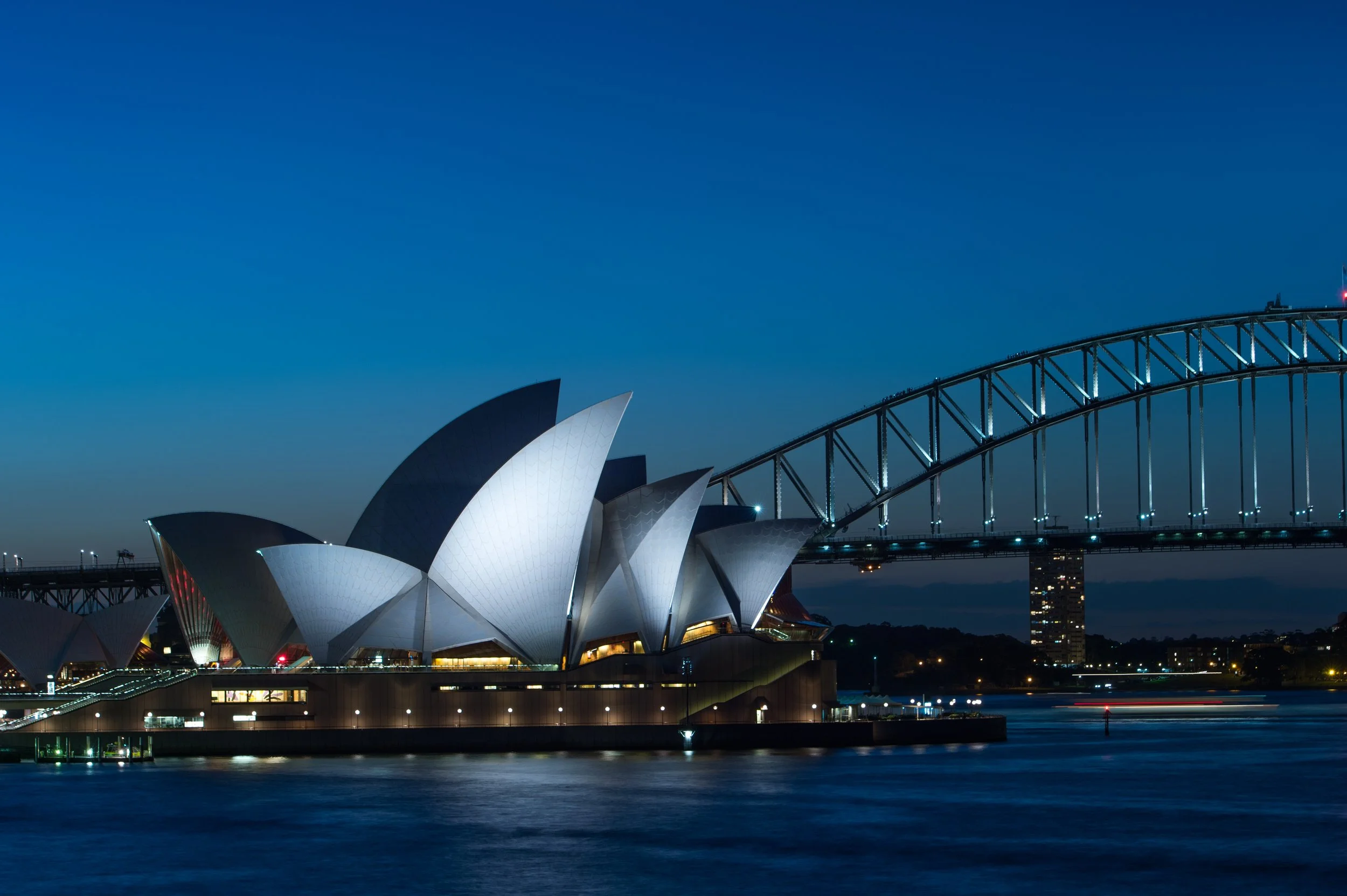 The Sydney Opera House illuminated against a dusk sky, with the Harbour Bridge adjacent, showing its arch and steel framework, along with water reflections below.