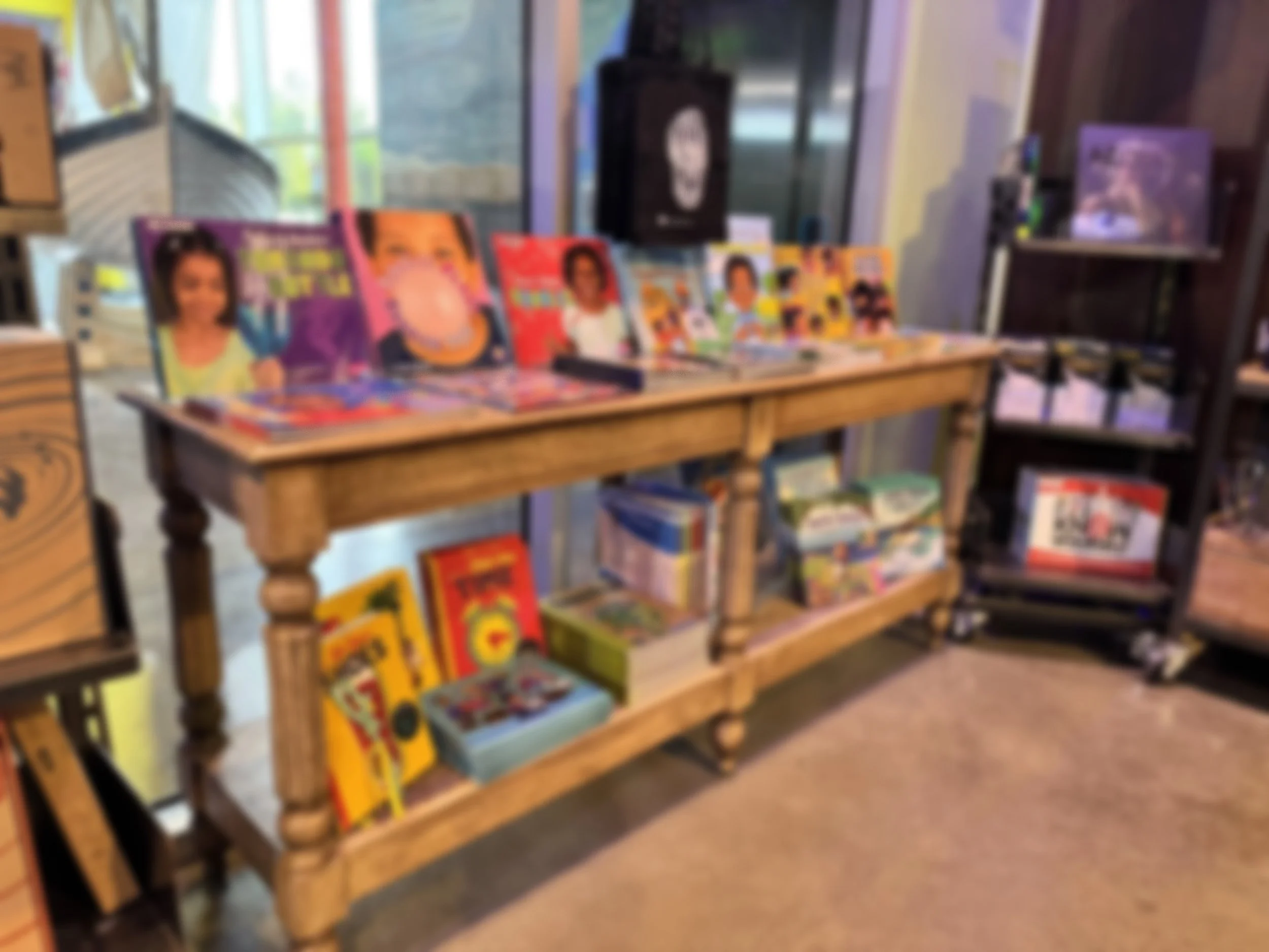 A wooden display table in a bookstore with colorful children's books on top and organized bookshelves underneath. In the background, part of a glass door and window are visible, letting in natural light.