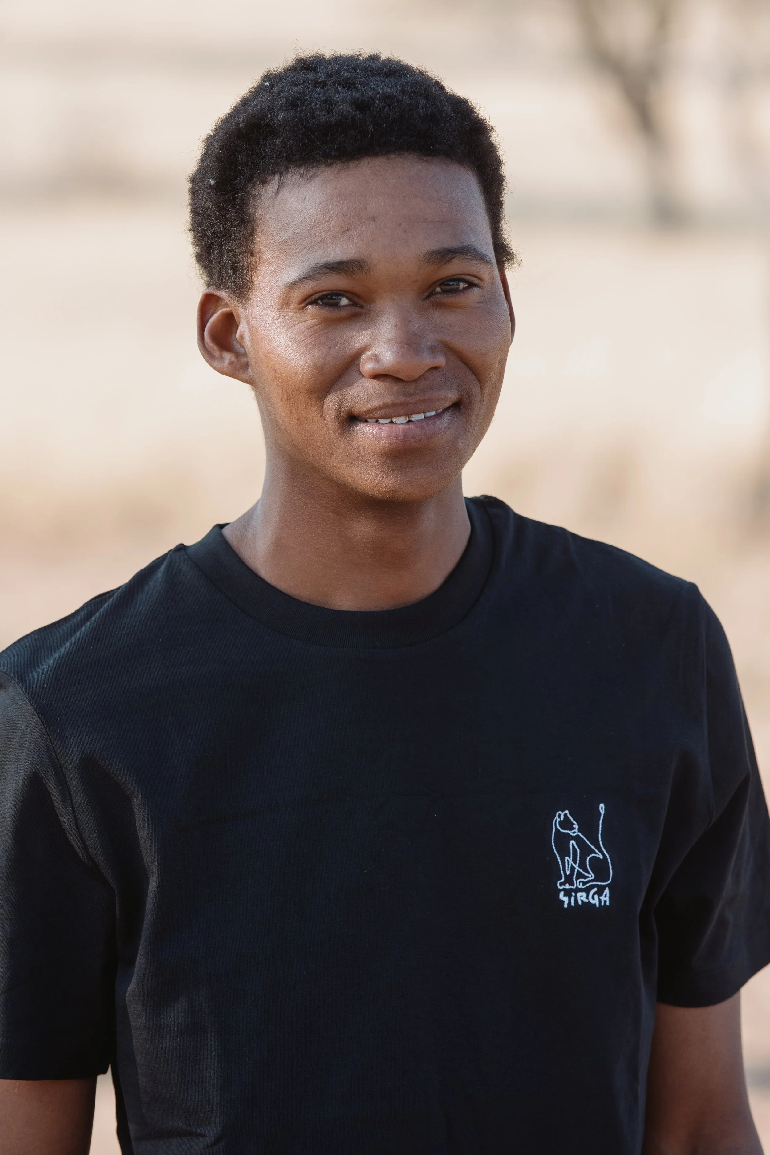 A person with short curly hair wearing a black T-shirt with a white logo of a lion and the word 'SIRGA' on it, standing outdoors with a blurry natural background.
