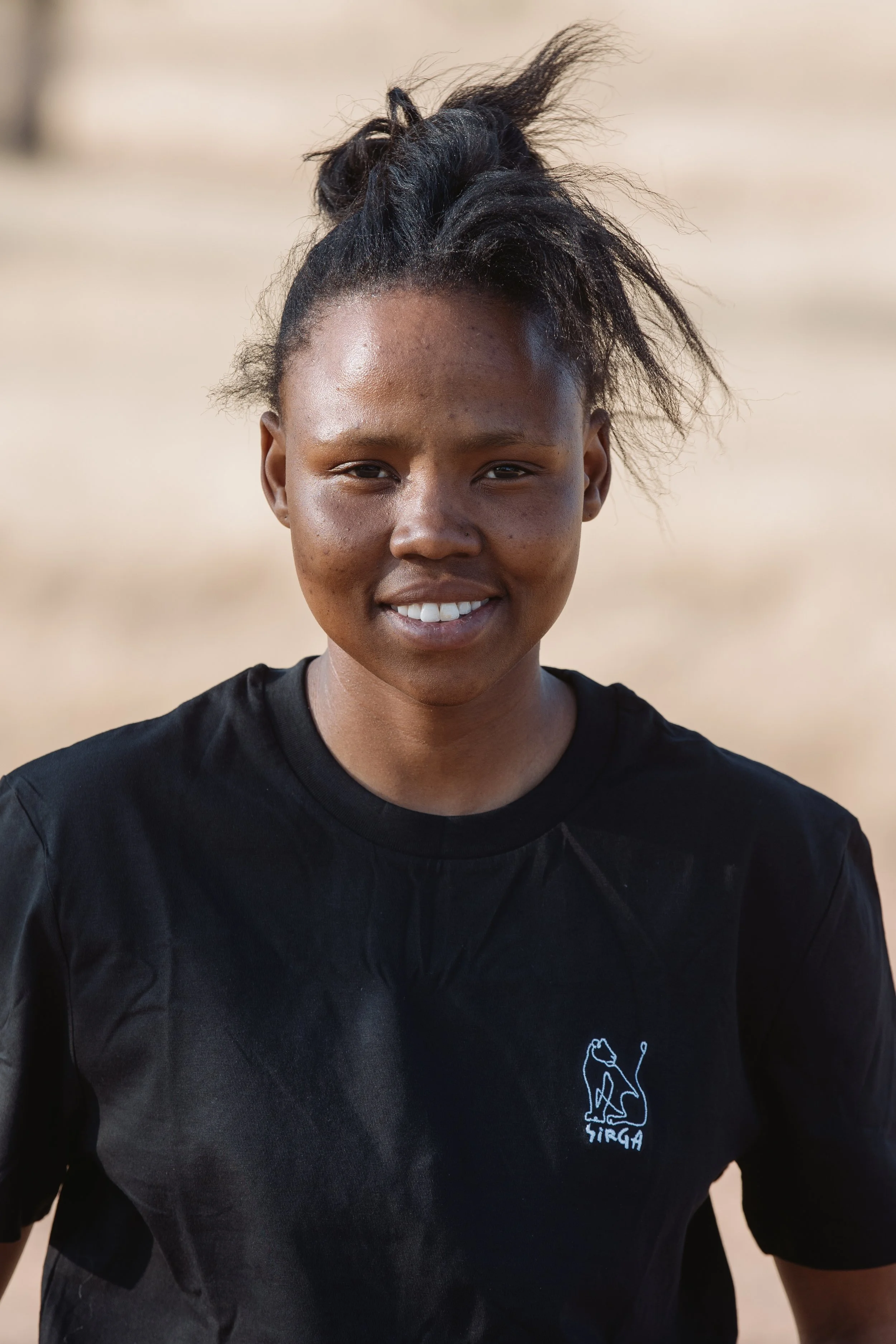 A young woman with dark skin and a black shirt smiling outdoors on a sunny day.