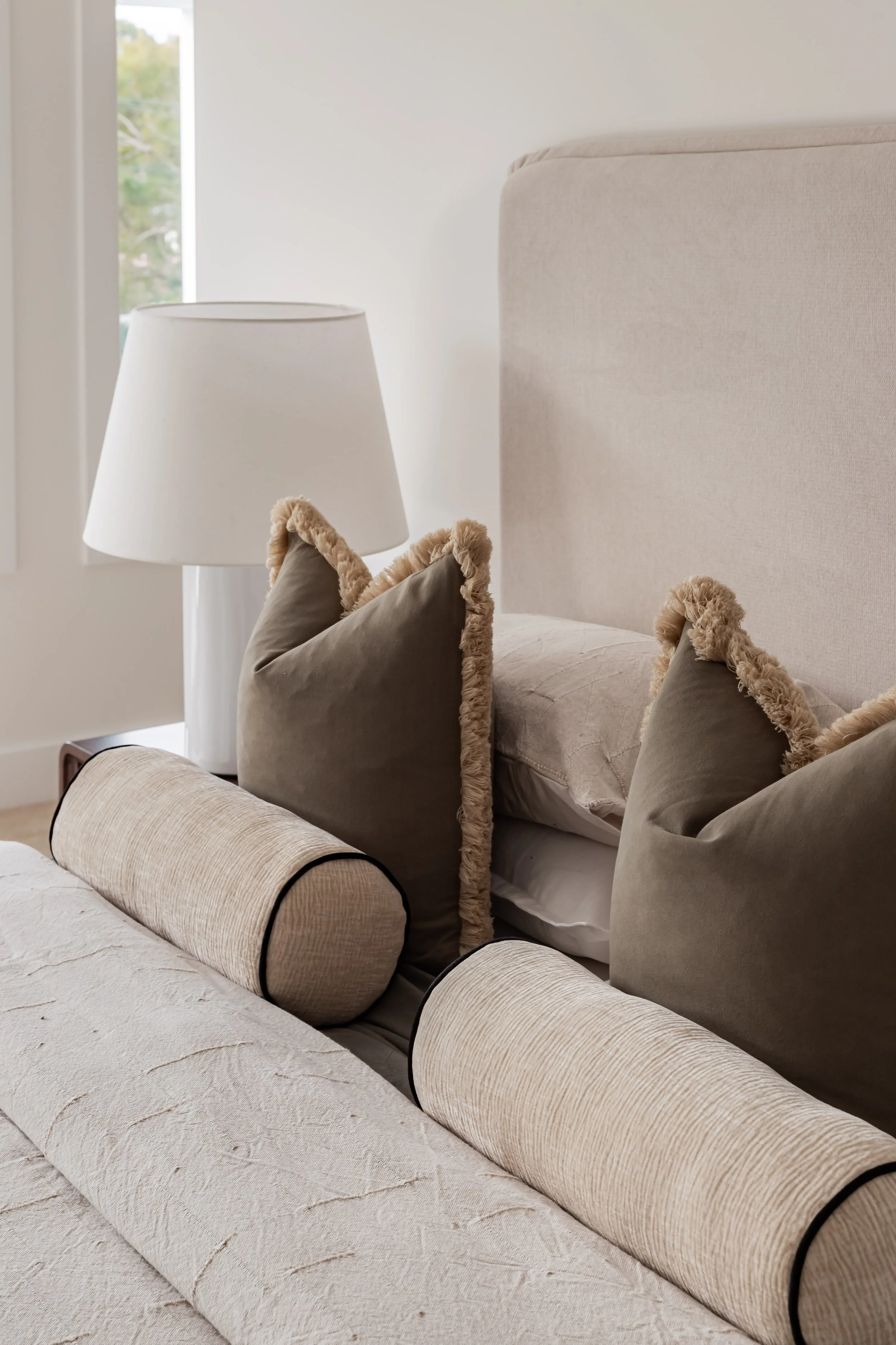 Modern bedroom with gray upholstered bed, white and gray pillows, a black nightstand with a large flower arrangement in a glass vase, books, and a window with a black frame, all illuminated by natural light.