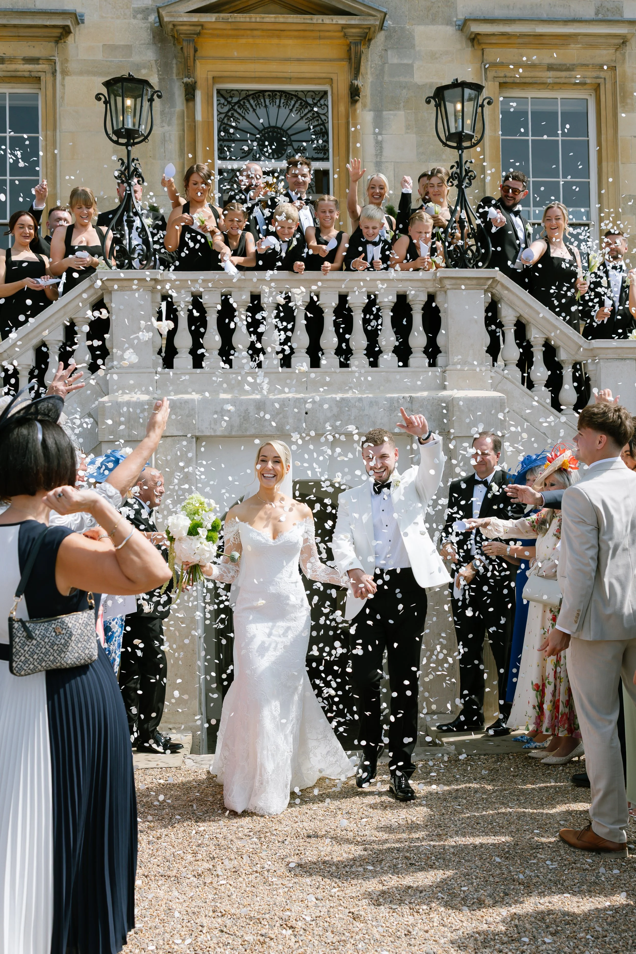 Newlyweds, the bride wearing a white lace gown and the groom in black trousers and a white tuxedo jacket, walk through white confetti being thrown by smiling wedding guests
